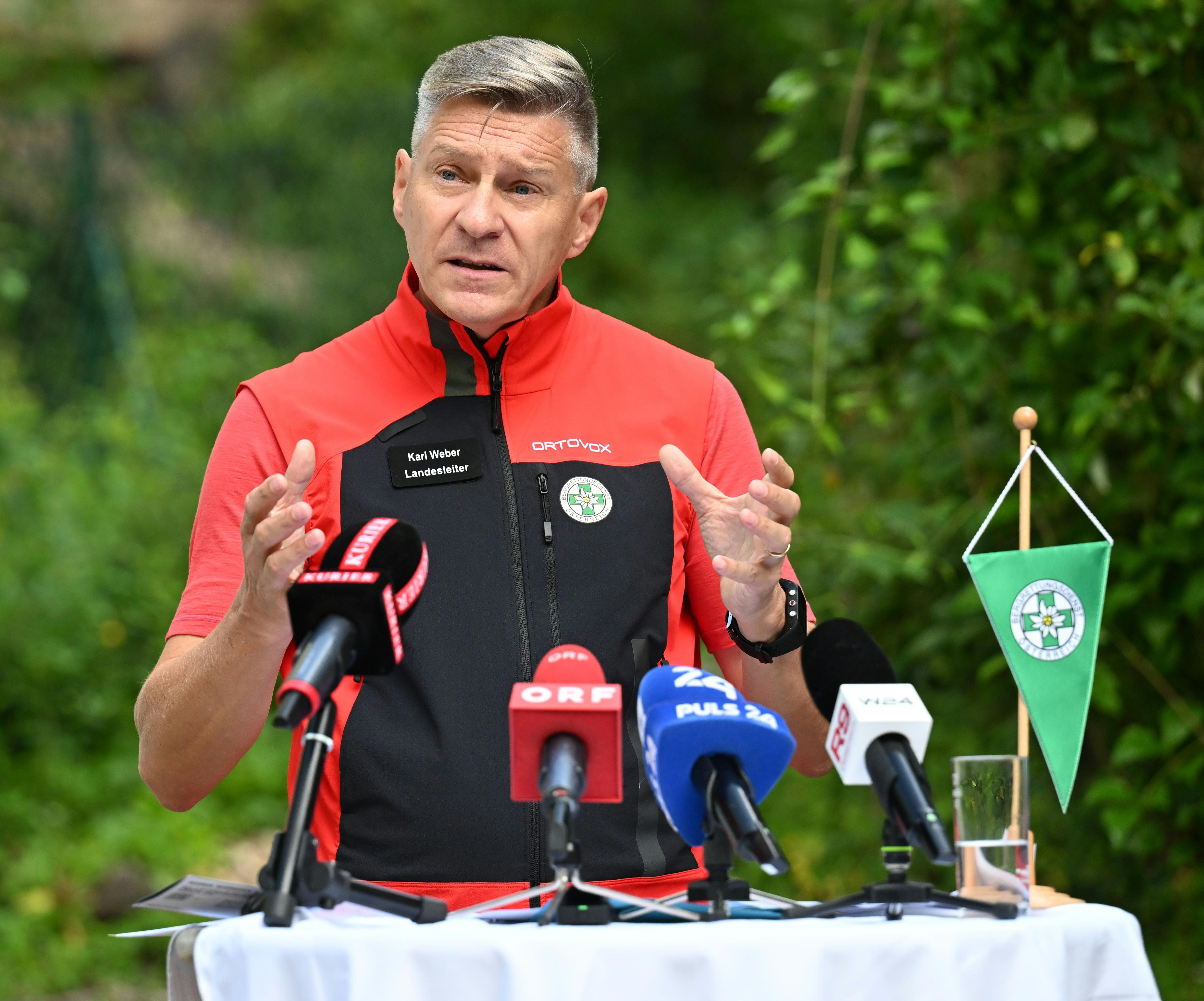 Ein Mann in roter Weste spricht bei einer Pressekonferenz im Freien vor mehreren Mikrofonen.