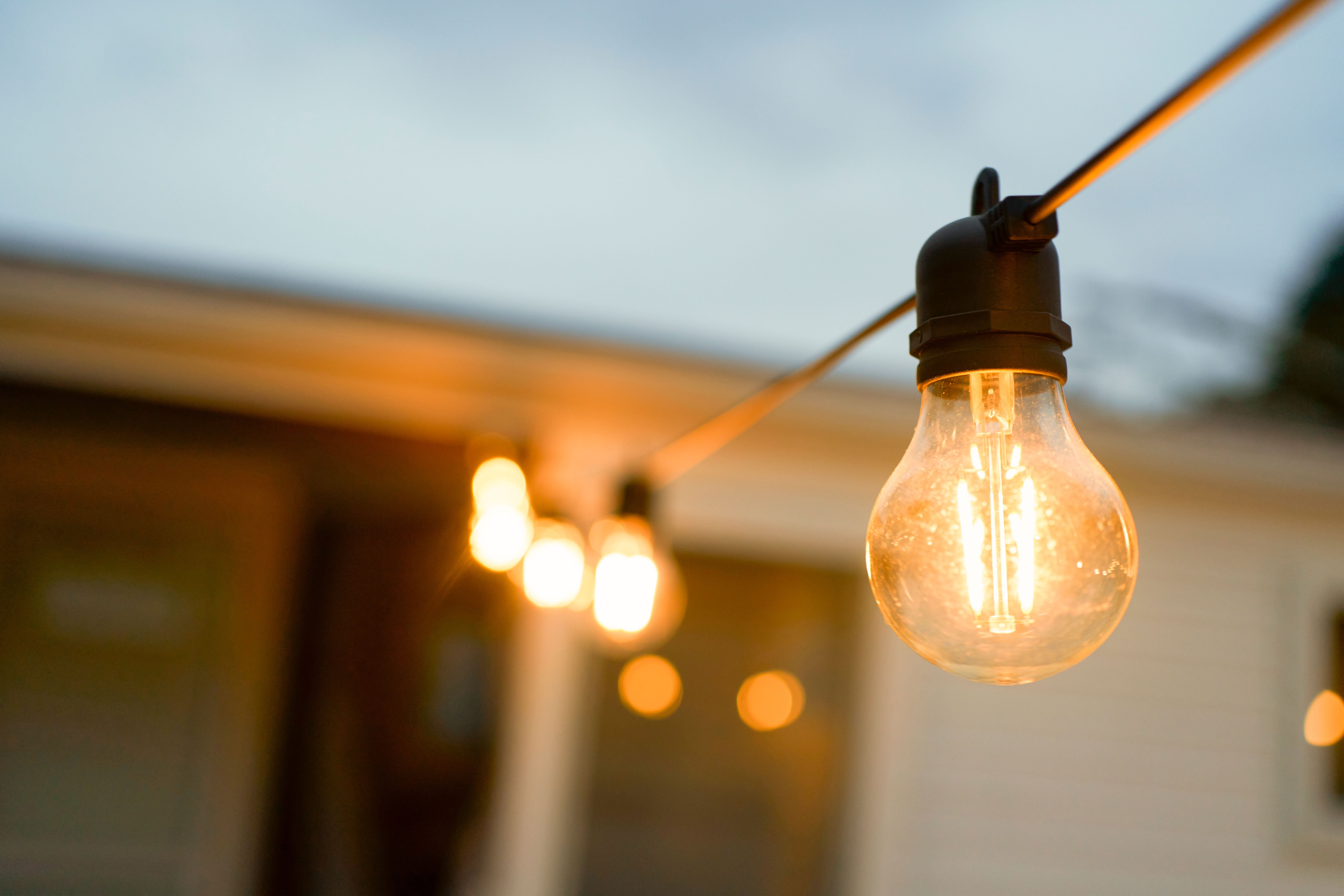 Eine Lichterkette mit warm leuchtenden Glühbirnen hängt vor einem unscharfen Haus.
