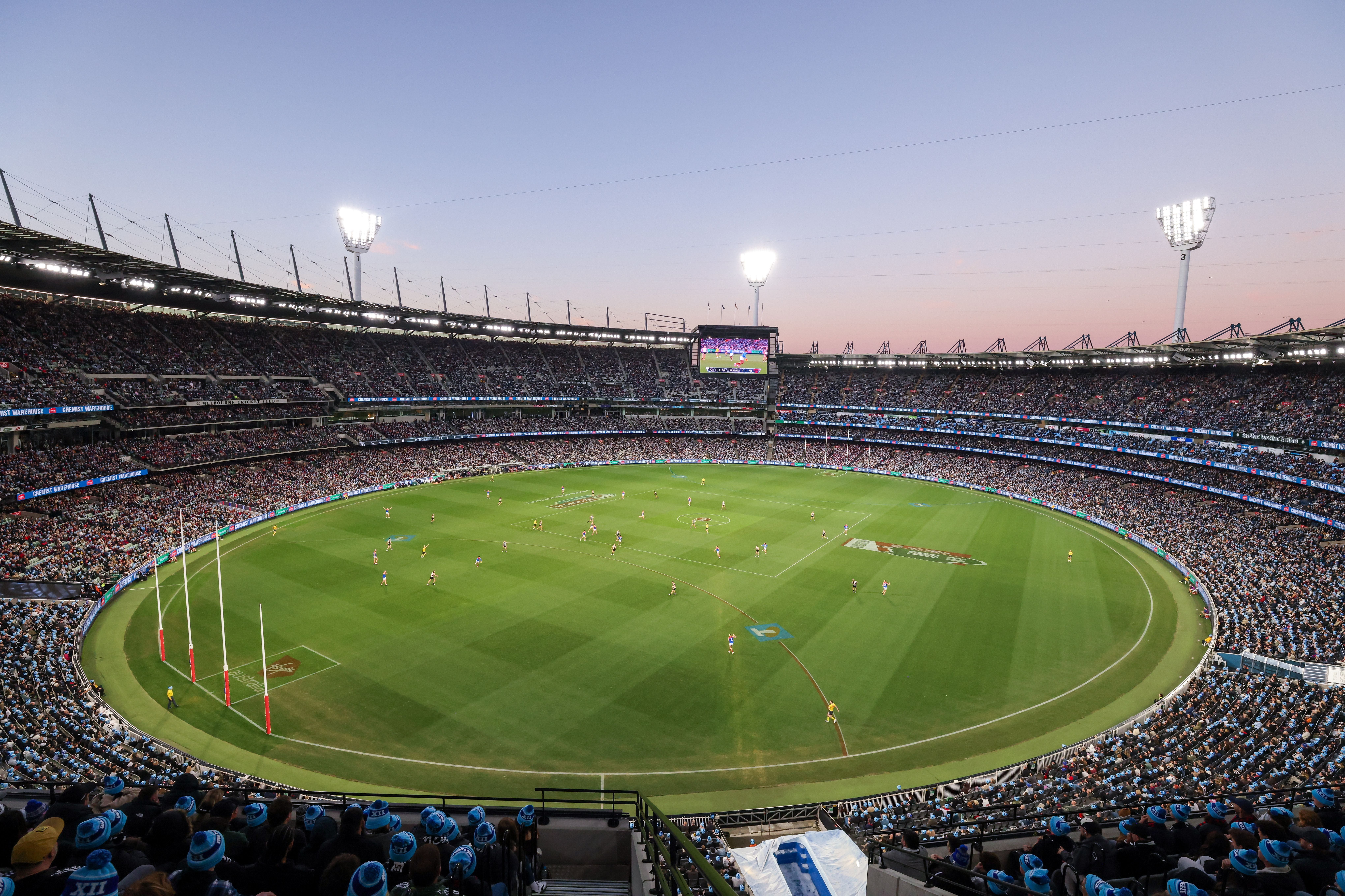 Großes, beleuchtetes Stadion in Melbourne mit Fußballspiel und vielen Zuschauern bei Sonnenuntergang.
