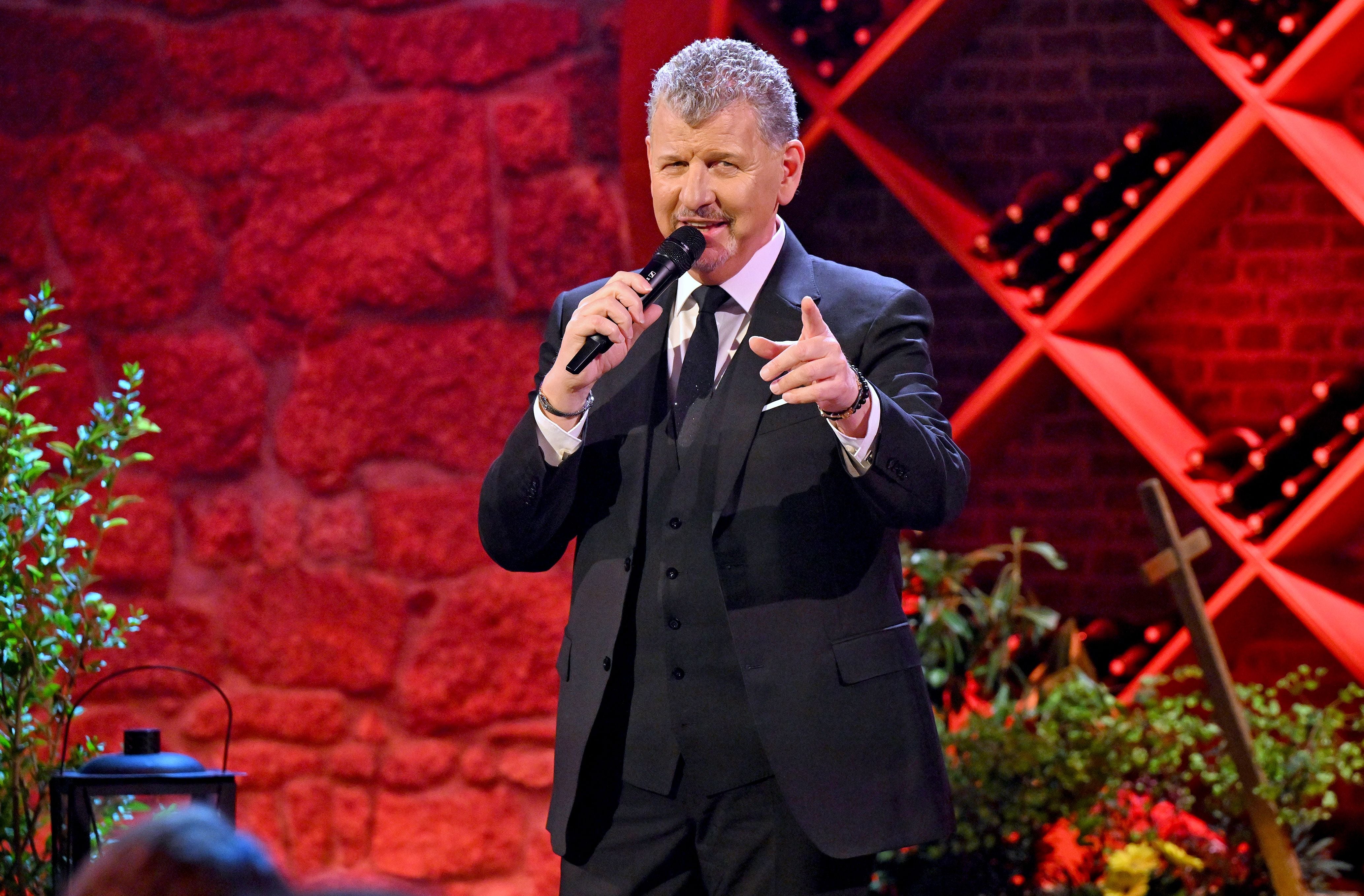 Ein Mann im Anzug singt mit Mikrofon auf einer Bühne mit rotem Hintergrund und Weindekor.