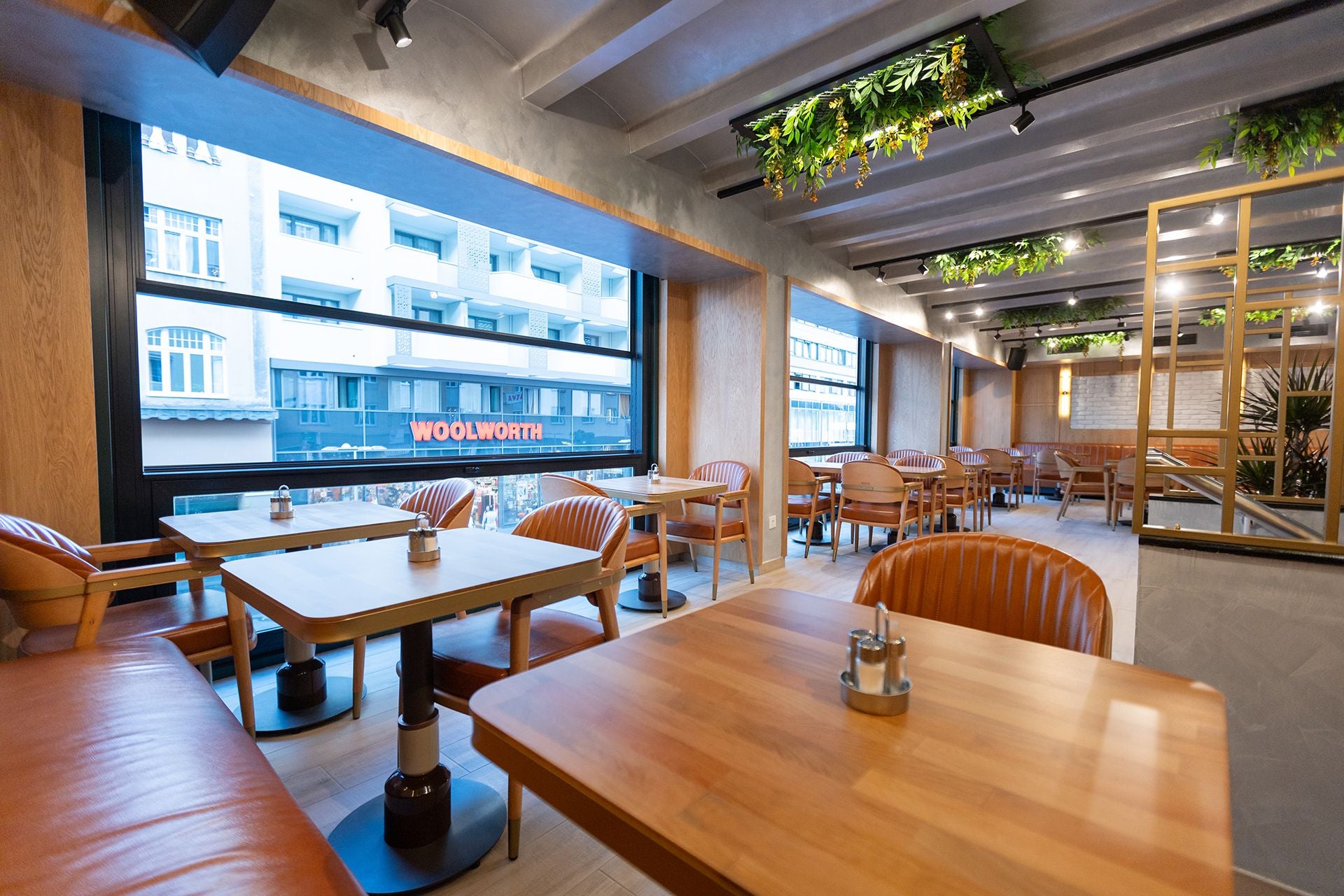 Modernes Café mit Holzmöbeln, großen Fenstern und begrüntem Deckenbereich, Blick auf Straße.