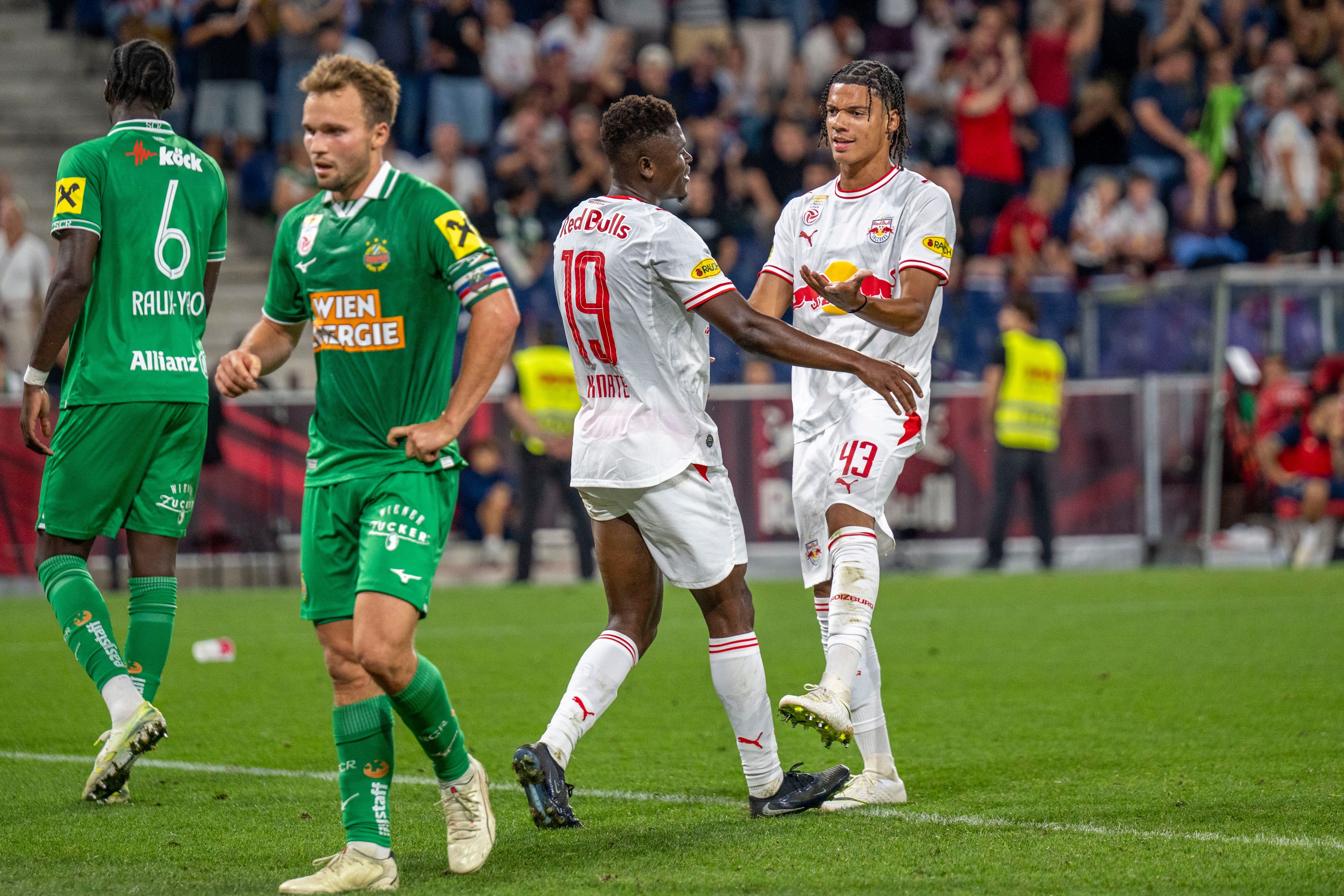 Zwei Spieler von Red Bull Salzburg jubeln auf dem Spielfeld, Rapid-Spieler gehen vorbei.