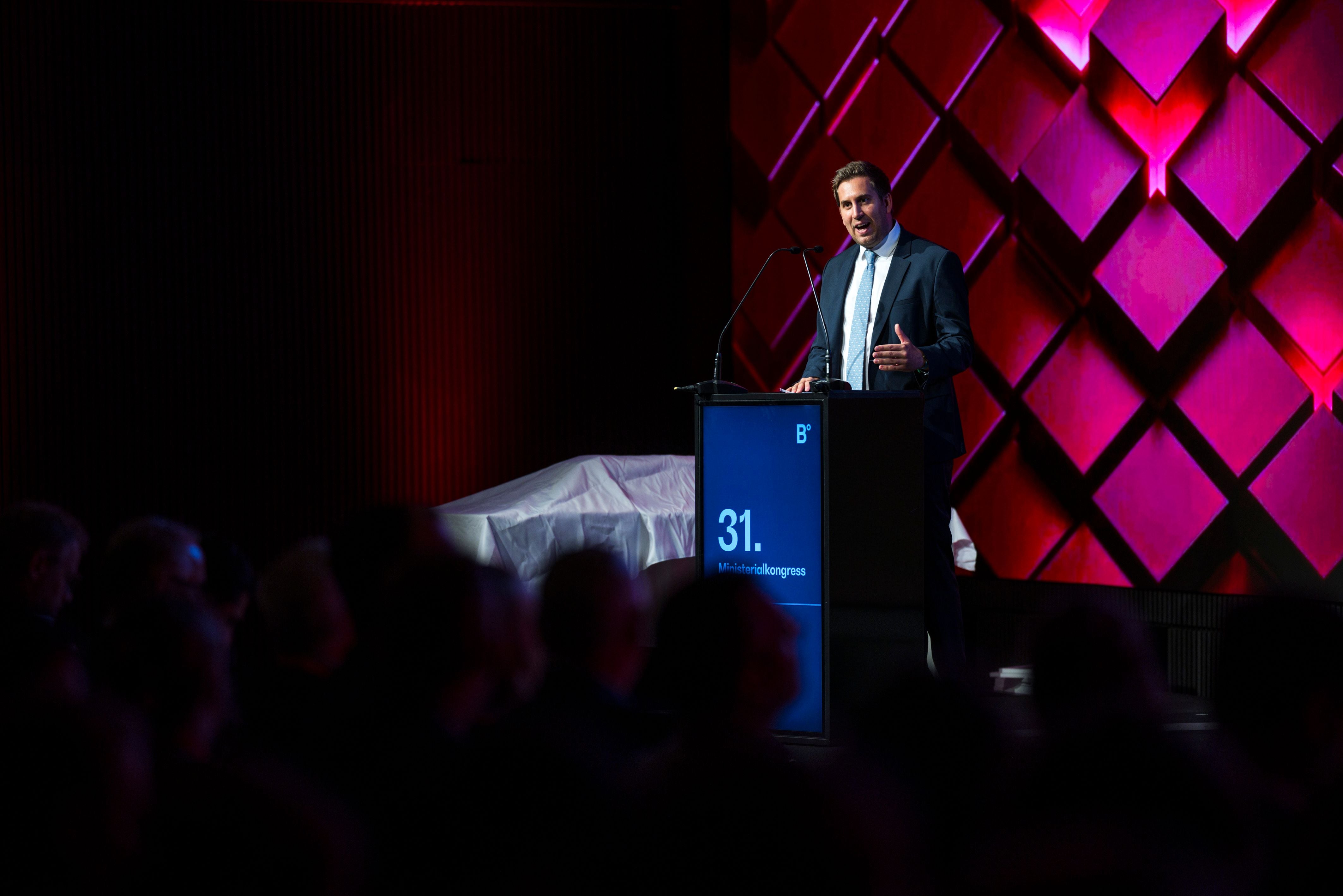 Ein Mann hält eine Rede am Rednerpult auf einem Kongress mit beleuchtetem Hintergrund.