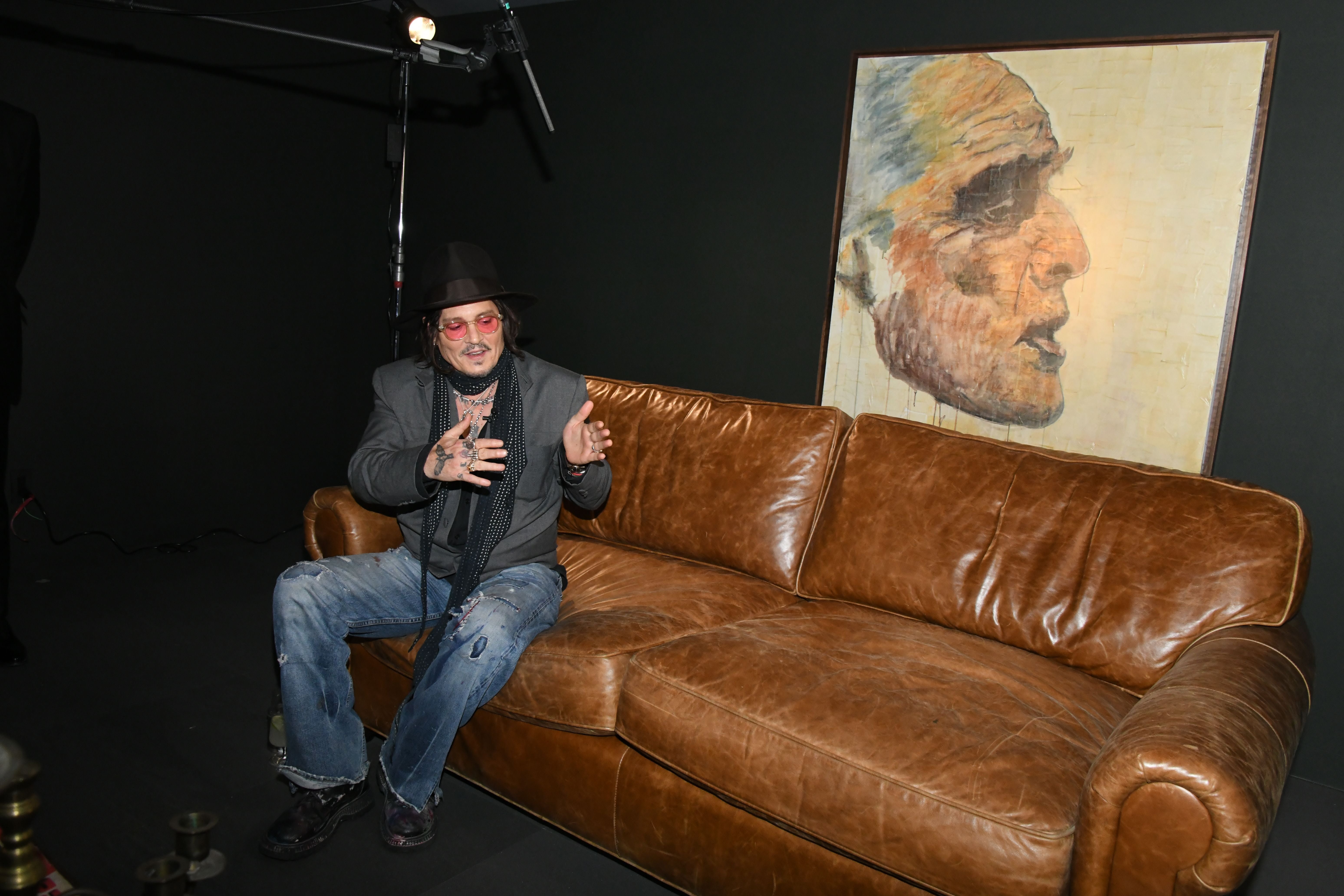 Mann mit Hut und Sonnenbrille sitzt auf braunem Ledersofa, großes Porträt im Hintergrund.