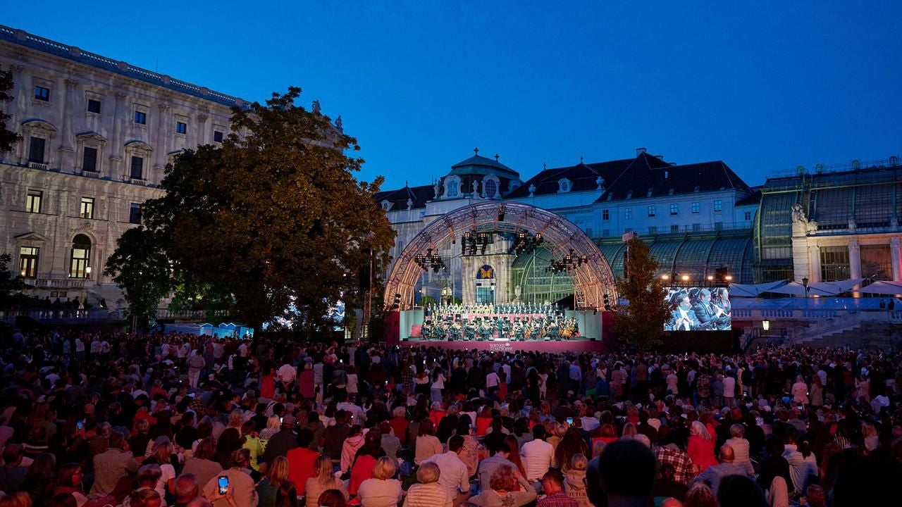 Großes Open-Air-Konzert auf einer Bühne vor historischem Gebäude bei Abenddämmerung, viele Zuschauer.