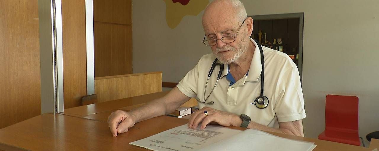 Ein älterer Mann mit Stethoskop liest ein Dokument an einem Holztresen.