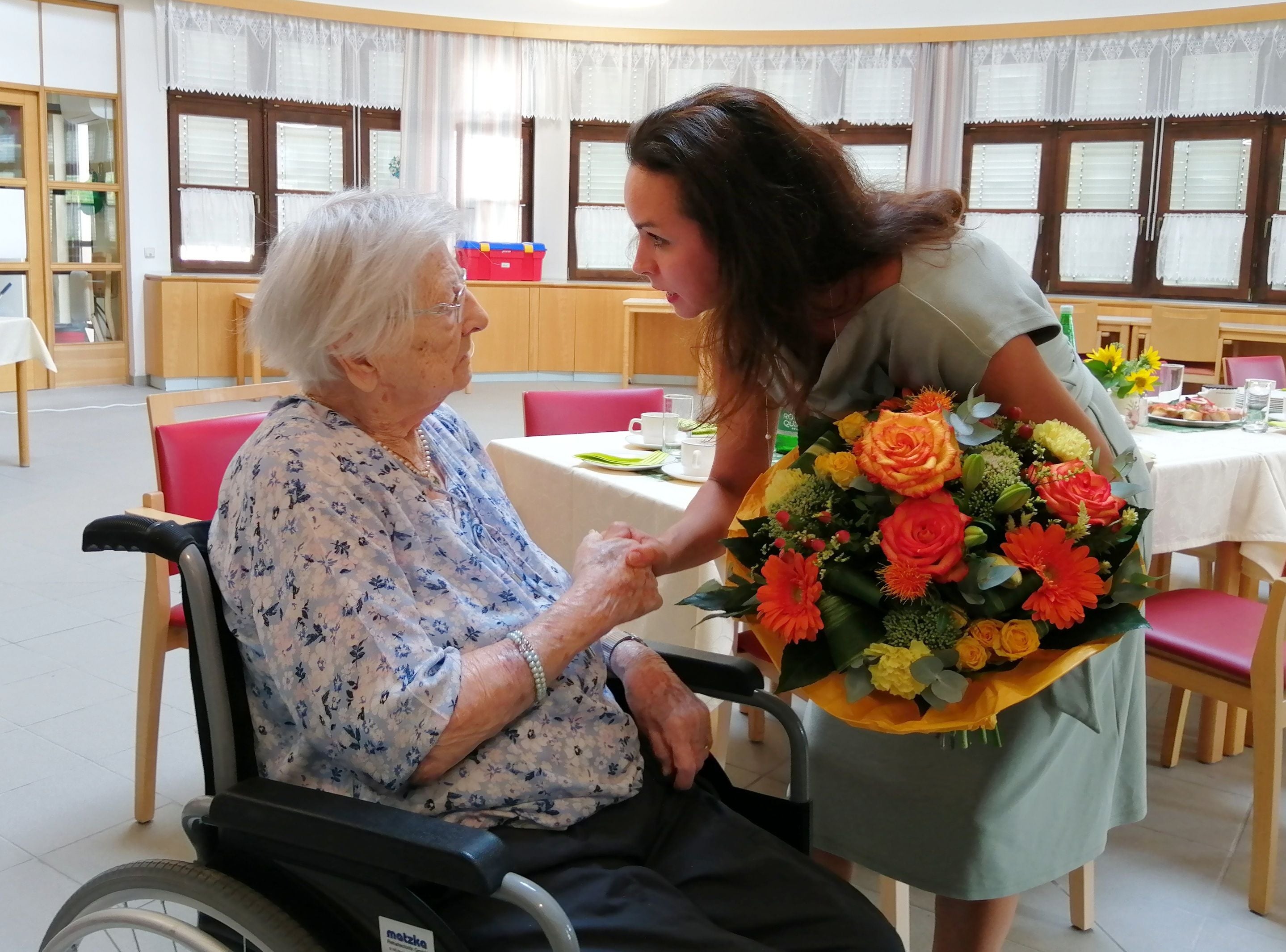 Ältere Frau im Rollstuhl bekommt zum Geburtstag Blumen von einer jüngeren Frau überreicht.