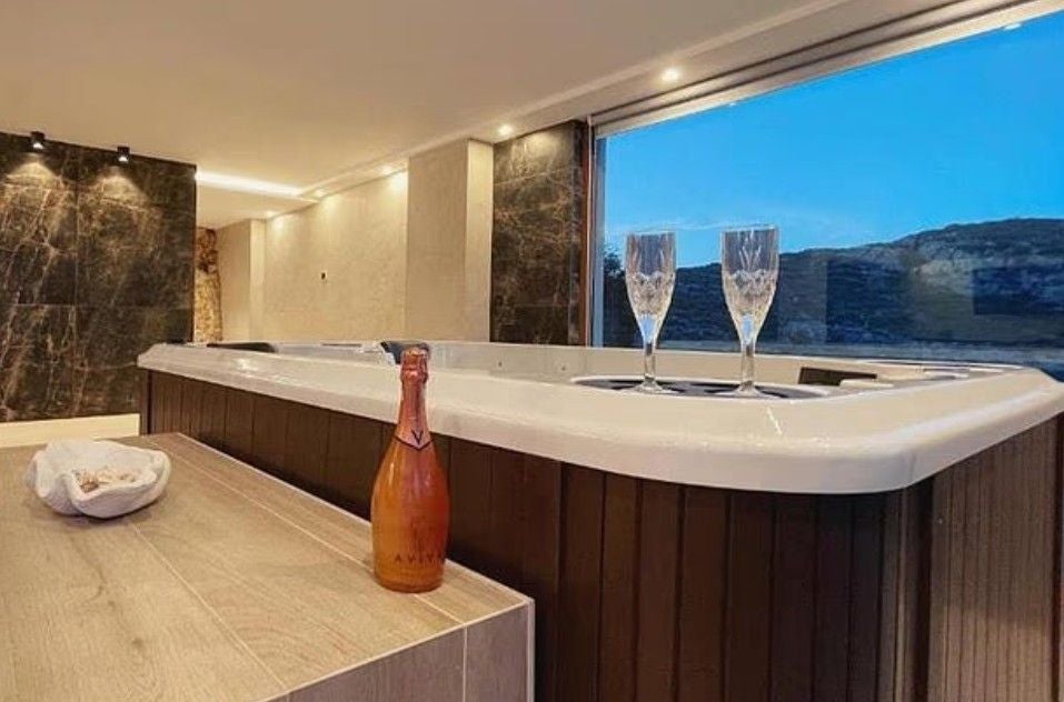 Whirlpool mit zwei Sektgläsern, Flasche und Blick auf Hügel durch großes Fenster.