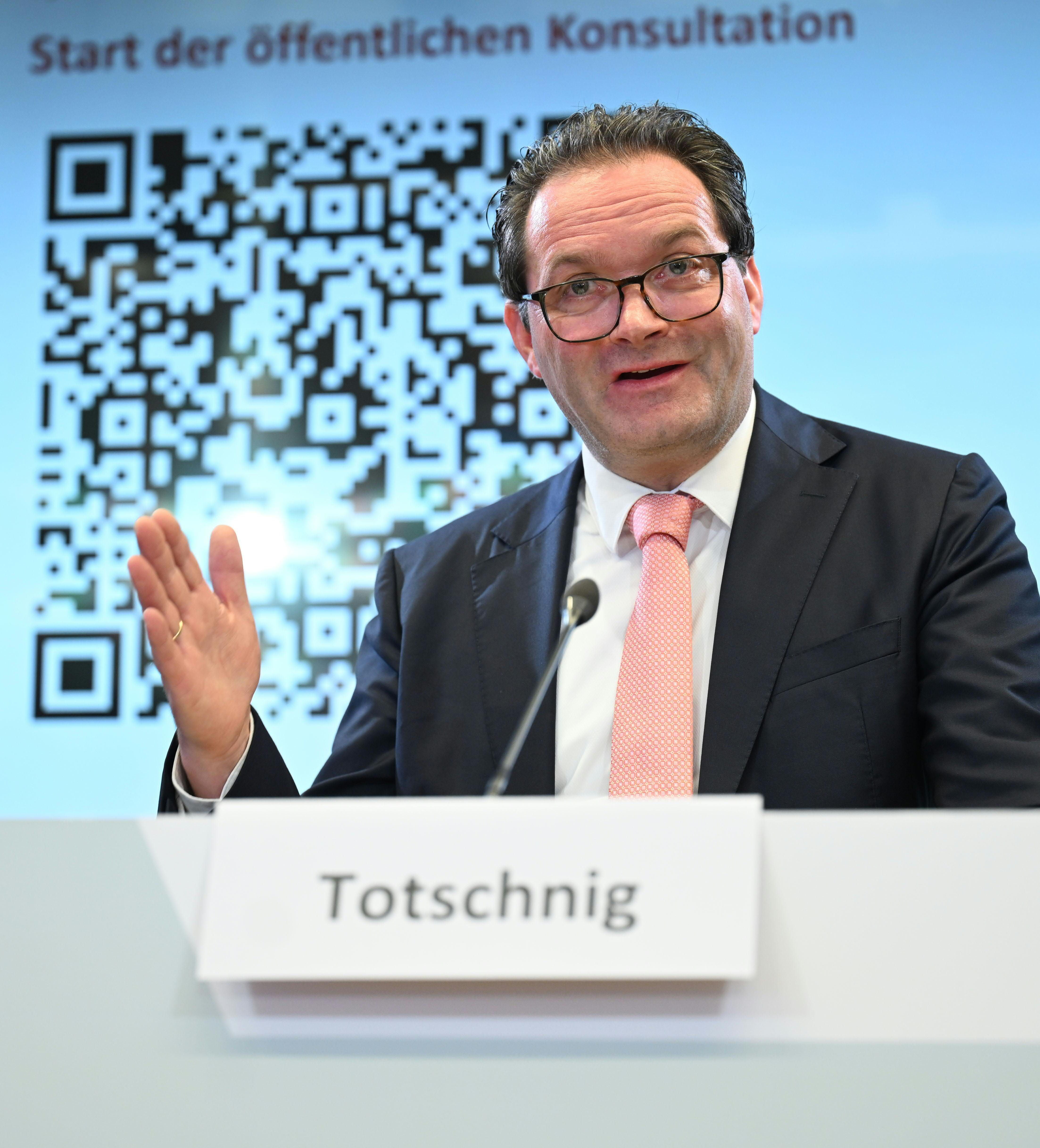 Ein Mann im Anzug spricht auf einer Pressekonferenz, hinter ihm ein QR-Code und ein Namensschild "Totschnig".