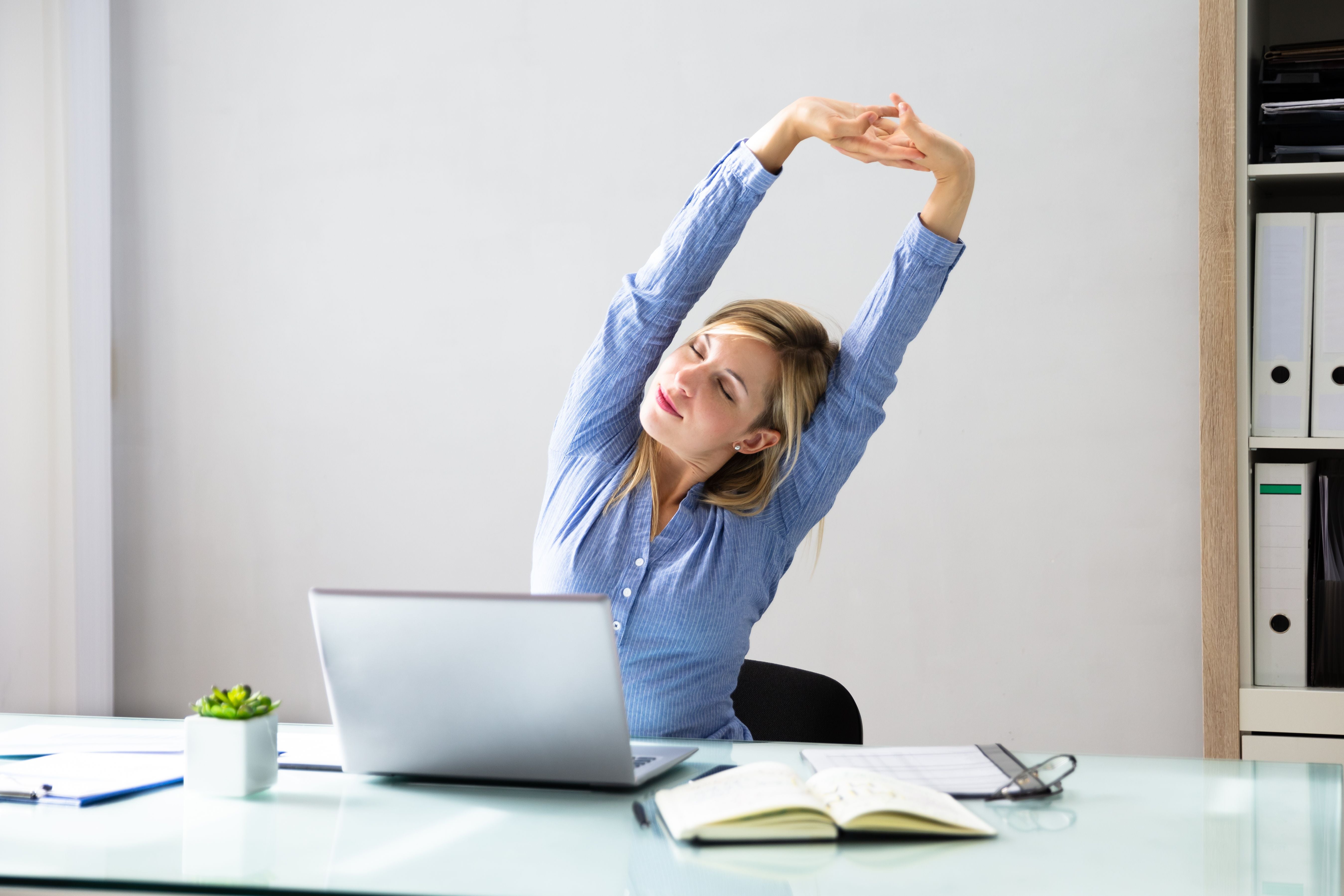 Junge Frau streckt sich am Schreibtisch vor ihrem Laptop im Büro.