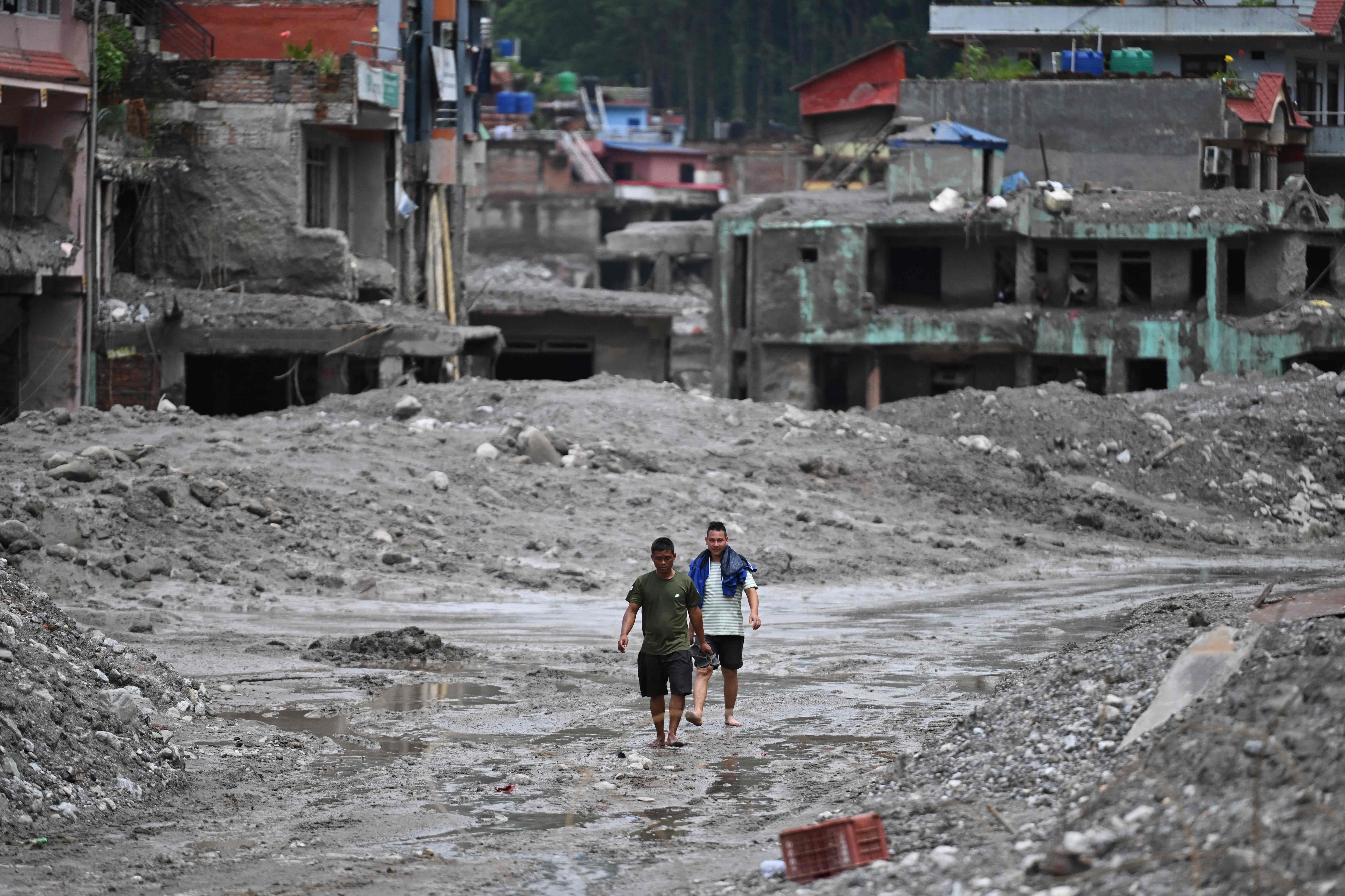 Zwei Männer gehen barfuß durch schlammüberschwemmte Straßen vor zerstörten Häusern in Nepal.