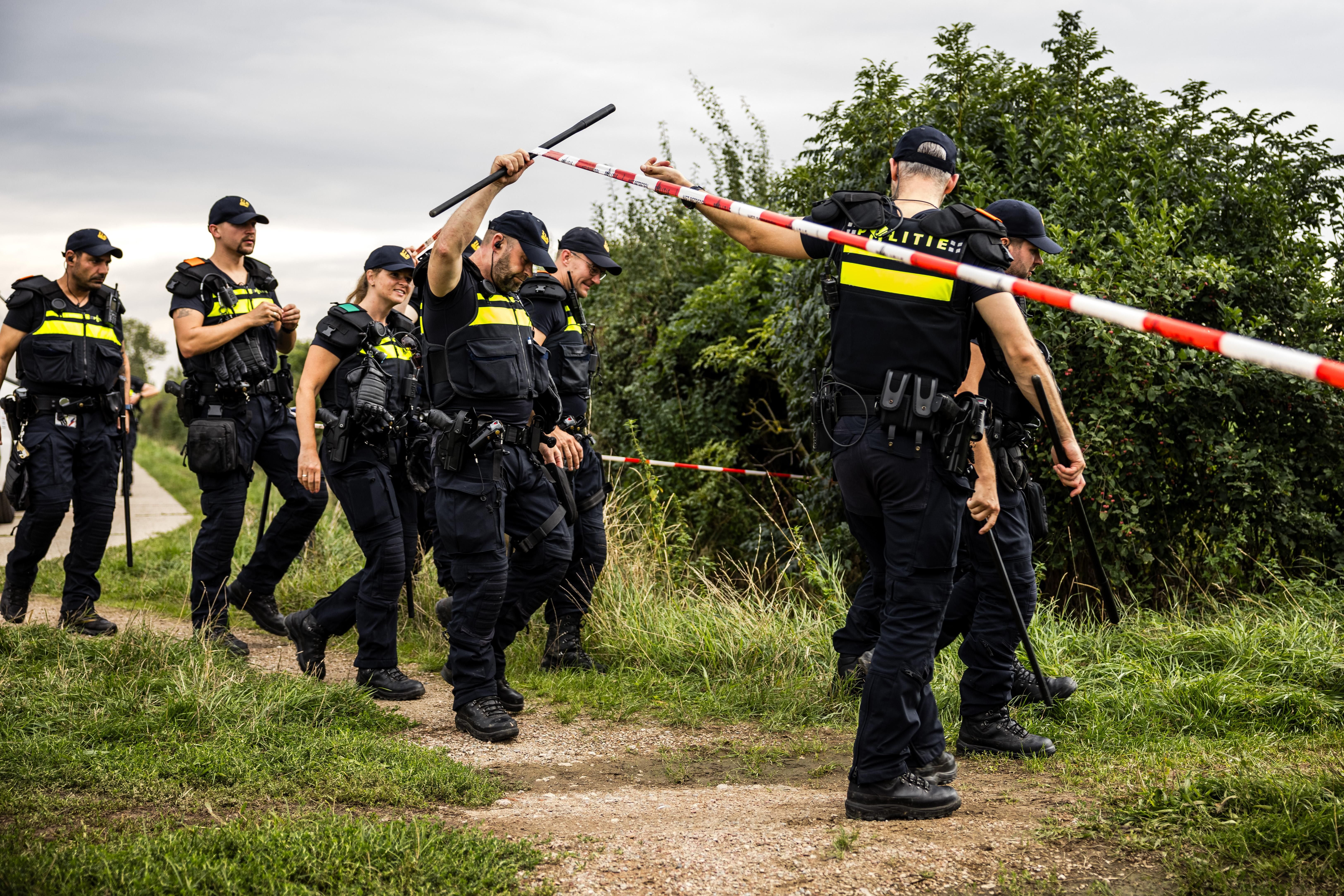 Polizisten durchsuchen ein Feld hinter einer Absperrung in der Nähe von Büschen.