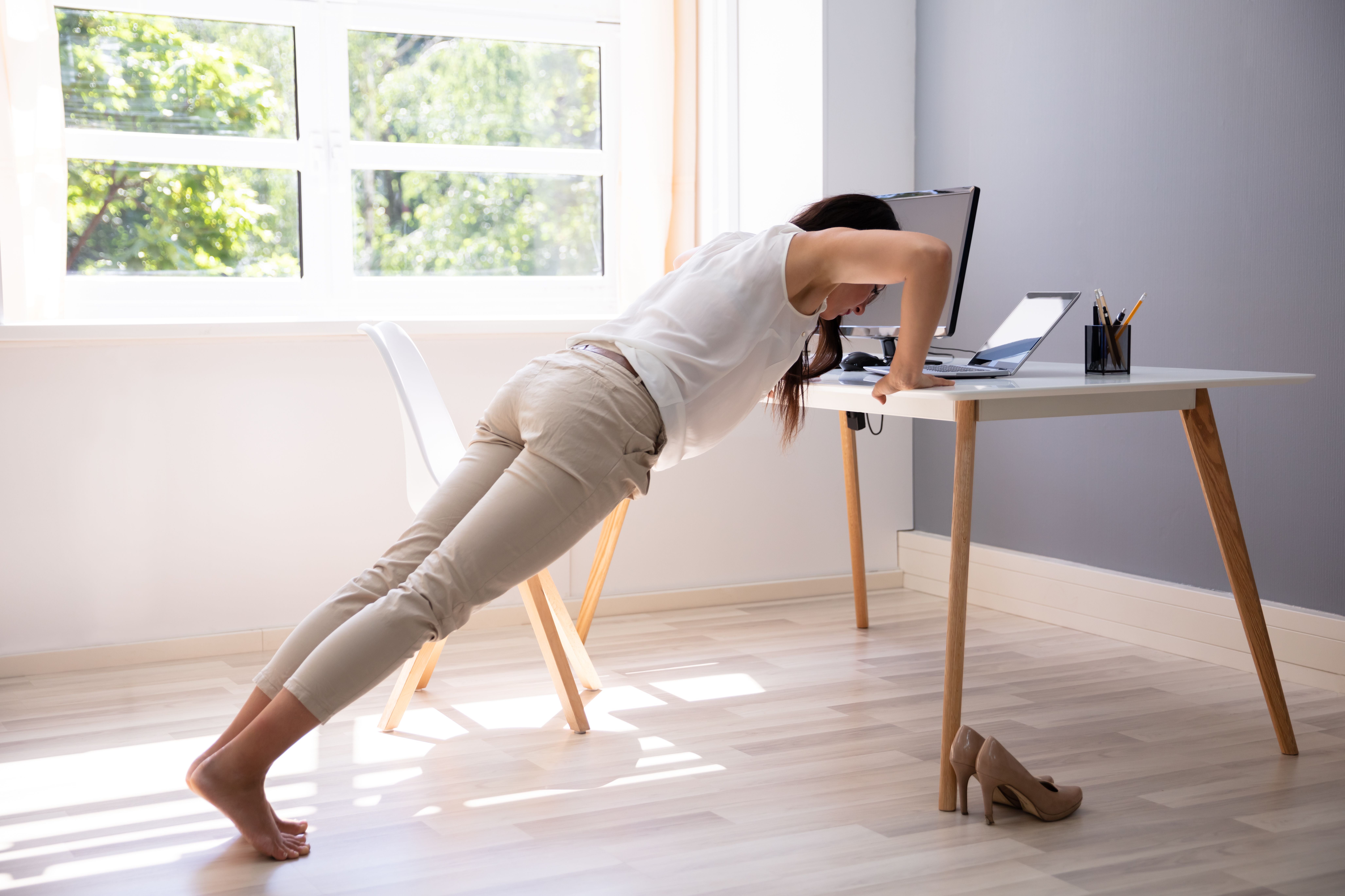 Frau macht am Schreibtisch im Büro gestützt Liegestütze, High Heels stehen auf dem Boden.