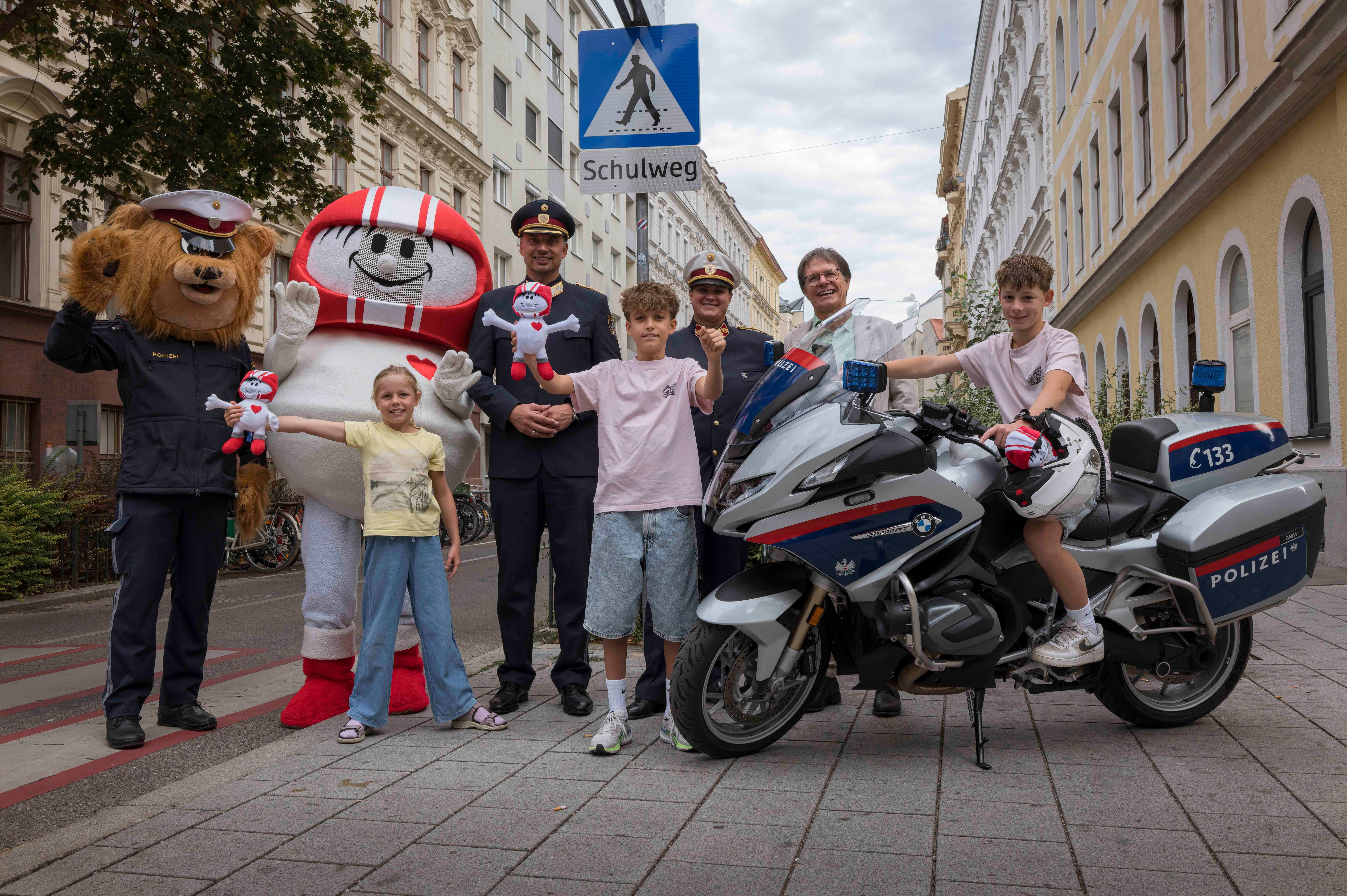 Kinder, Polizisten und Maskottchen posieren mit Polizeimotorrad unter einem "Schulweg"-Schild in Wien.