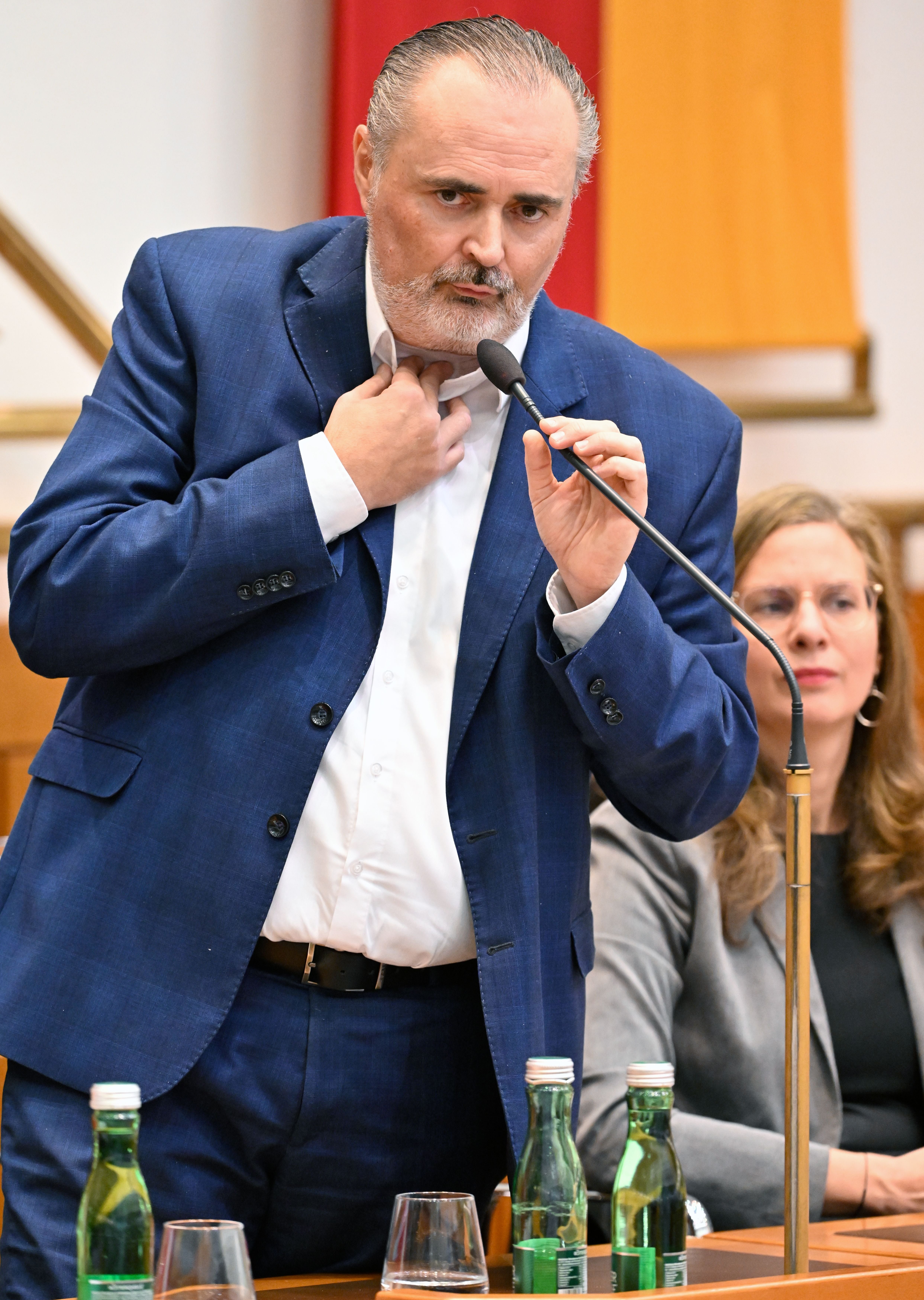 Ein Mann im blauen Anzug spricht während einer Sitzung an ein Mikrofon, vor ihm stehen Wasserflaschen.