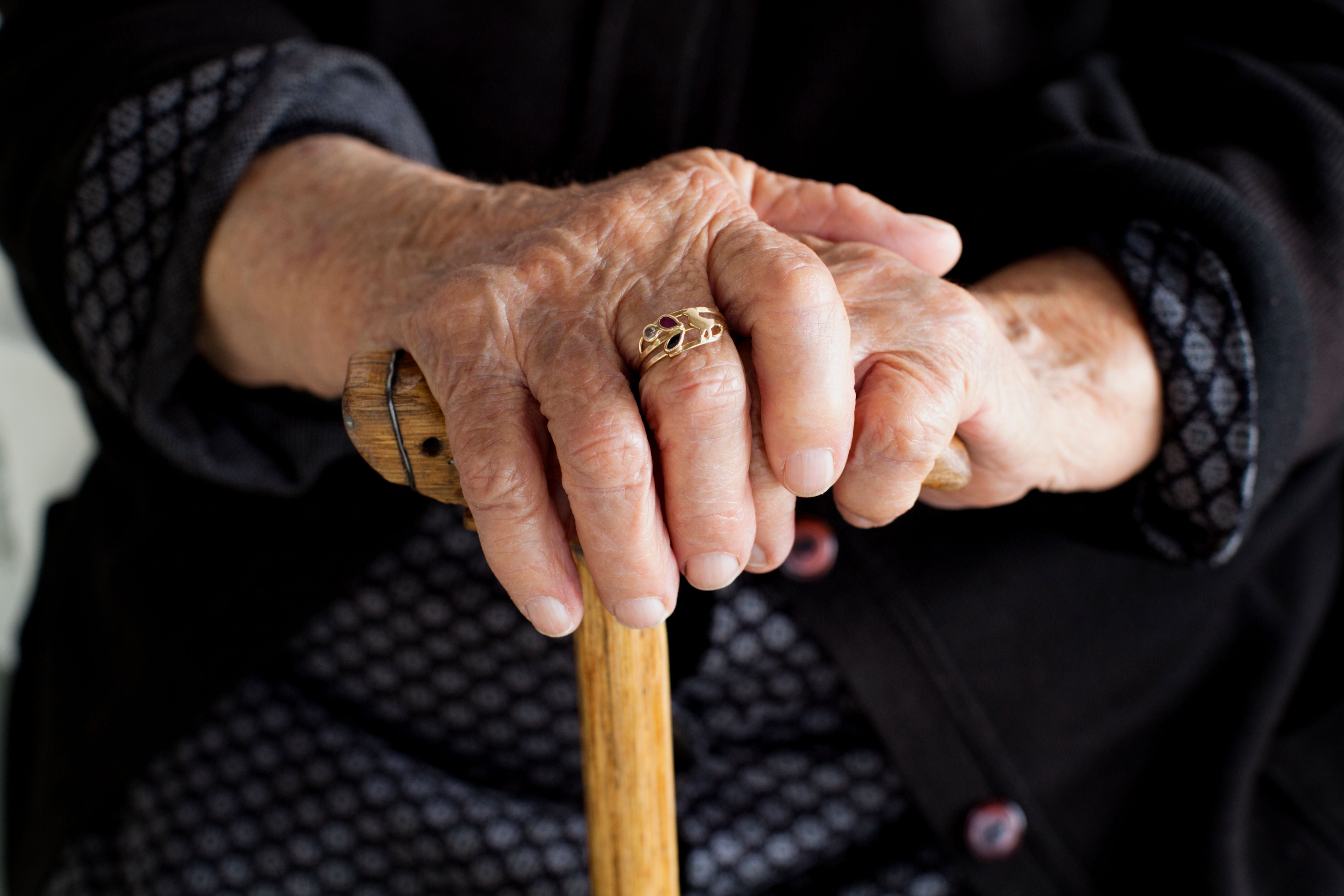 Faltenreiche Hände einer älteren Frau halten einen Holstock, ein goldener Ring ziert einen Finger.