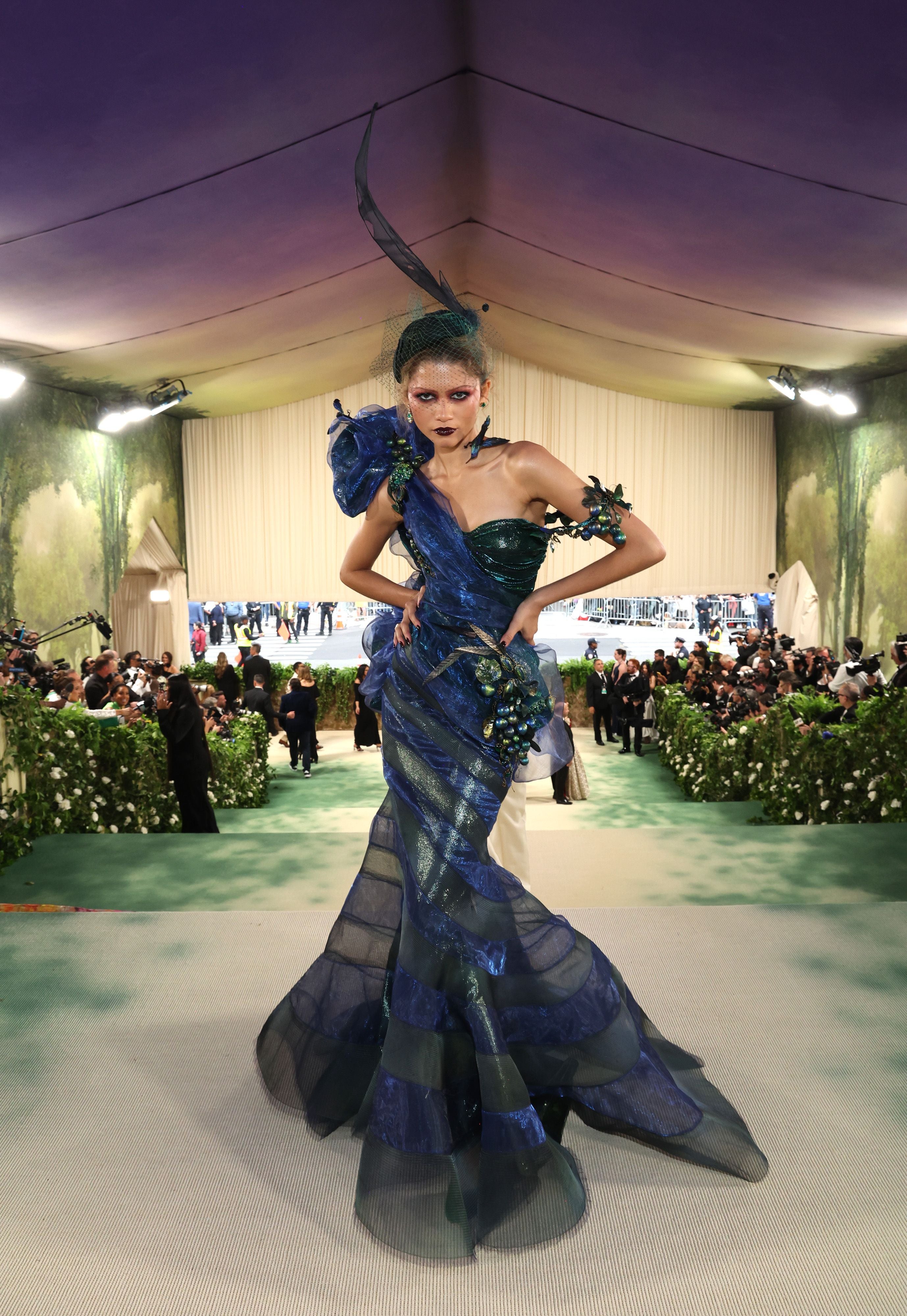 Frau in extravagantem blau-grünem Kleid mit Kopfschmuck auf grünem Teppich bei einer Gala.