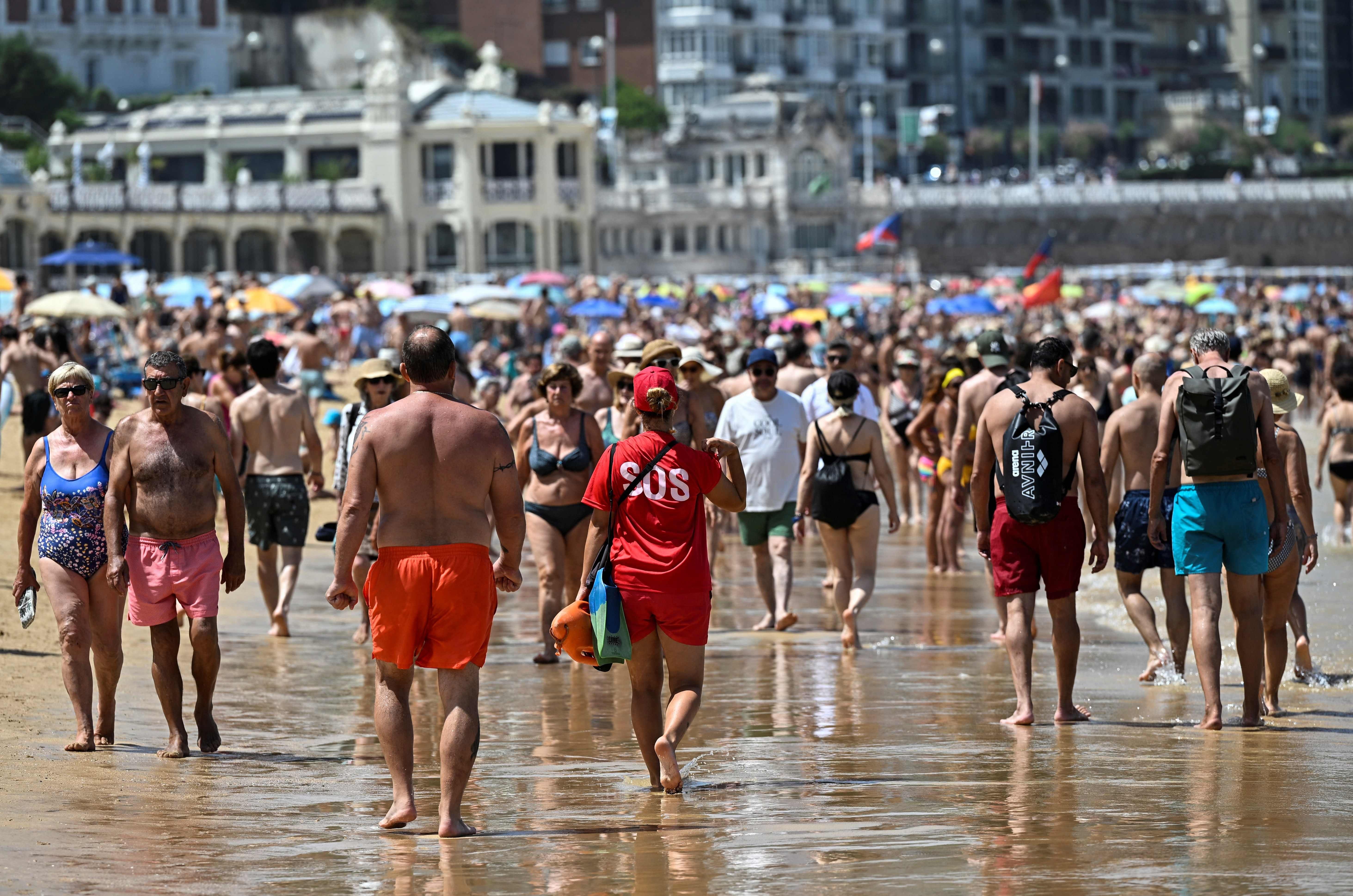 Menschenmengen spazieren an einem überfüllten Strand bei sonnigem Wetter; ein Rettungsschwimmer im Vordergrund.
