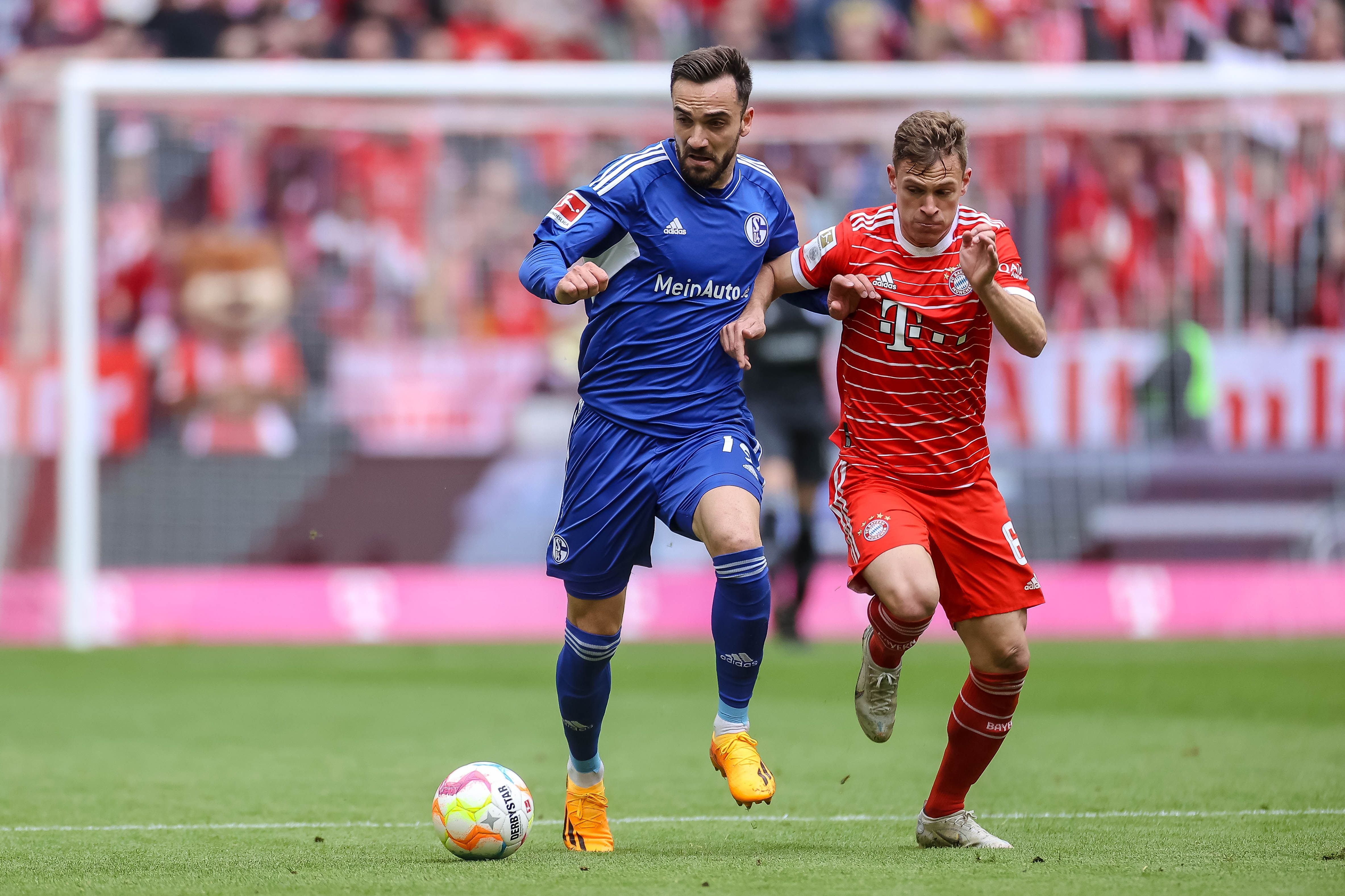Kenan Karaman im blauen Schalke-Trikot und Joshua Kimmich im roten Bayern-Trikot kämpfen auf dem Fußballplatz um den Ball.