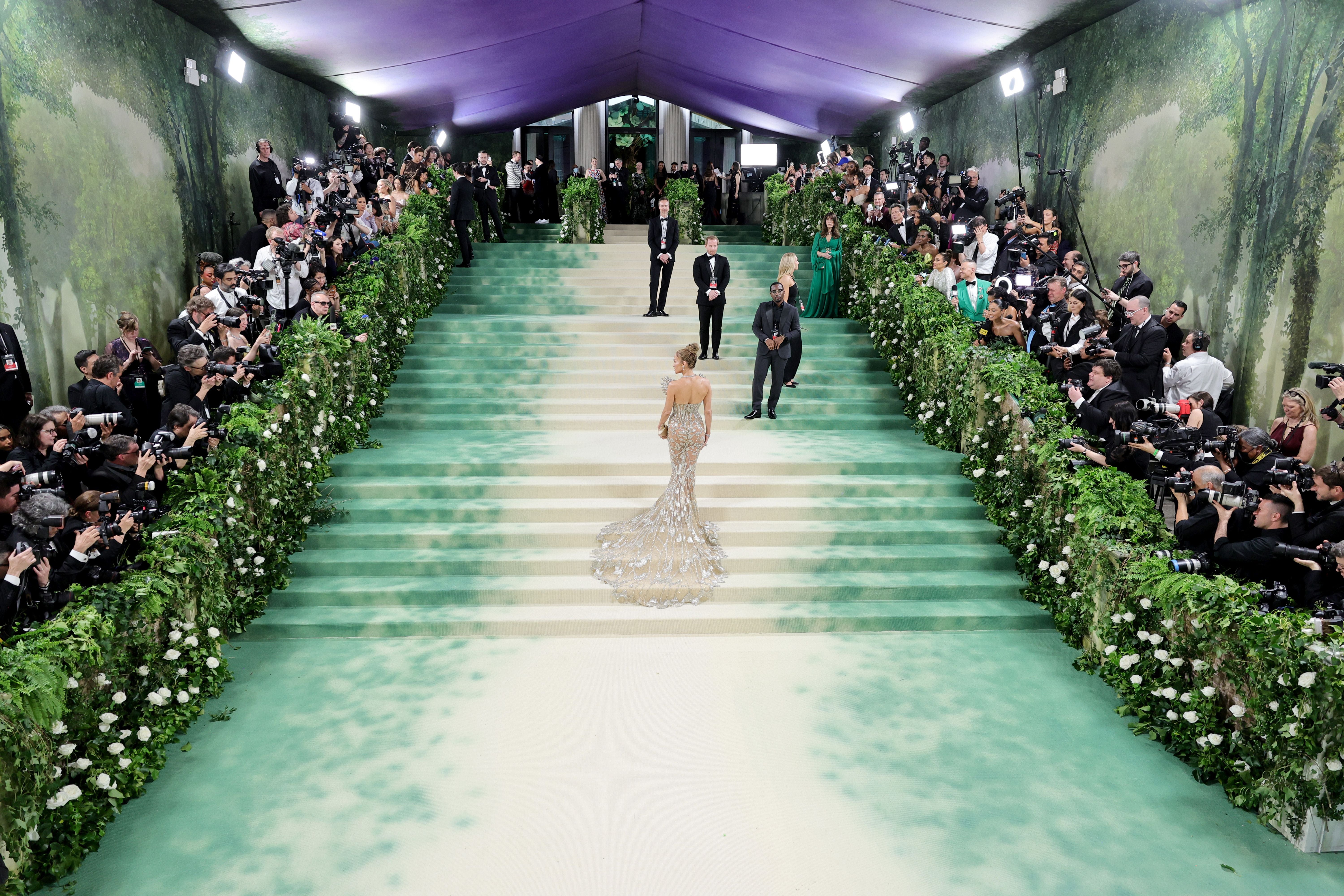 Eine Frau im glitzernden Kleid schreitet eine große, verzierte Treppe hinauf, umgeben von Fotografen.