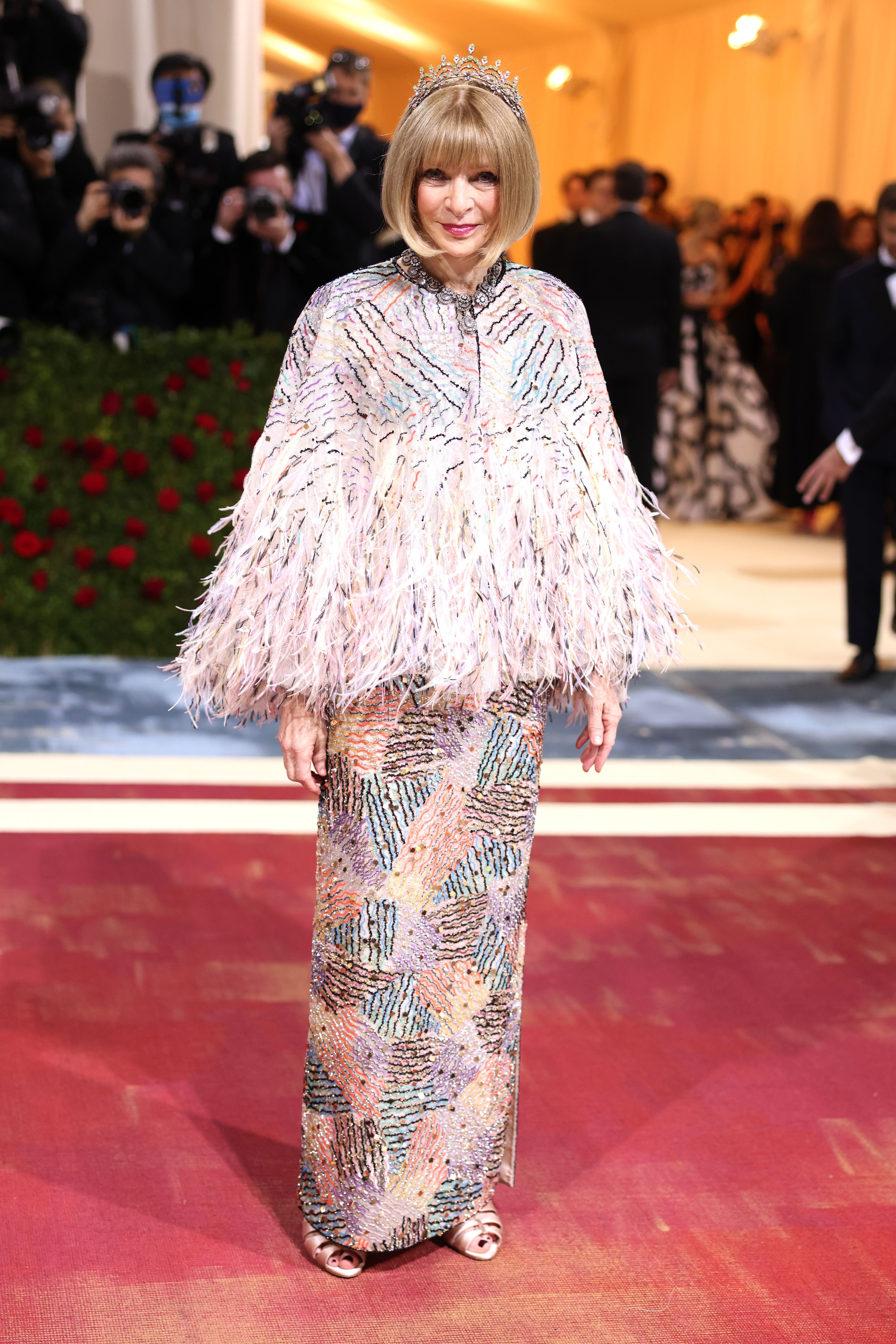 Frau mit Federumhang, langer Musterrobe und Krone auf rotem Teppich eines Gala-Events.