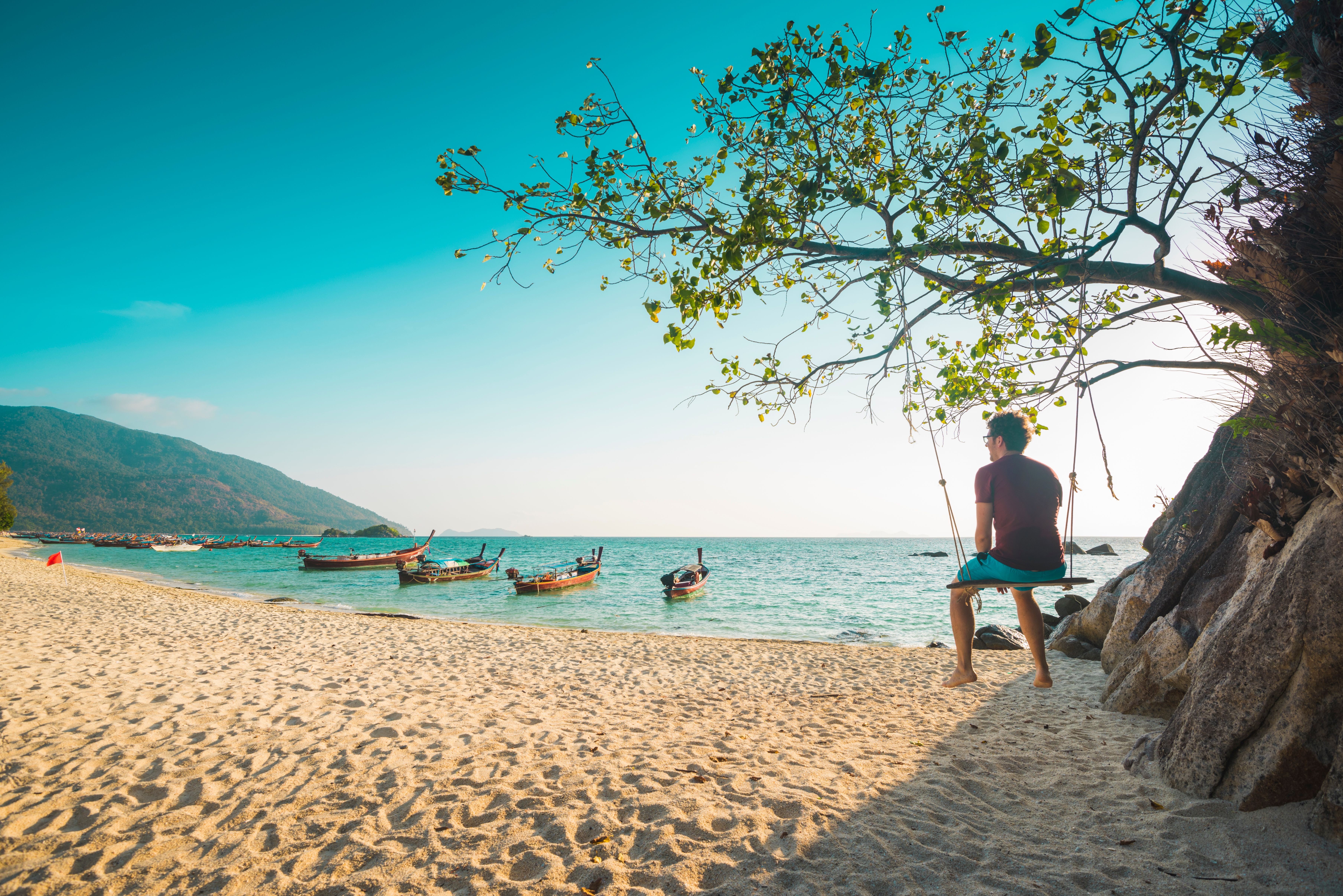 Mann sitzt auf einer Schaukel am Strand, Blick auf das Meer mit Booten und Bergen im Hintergrund.