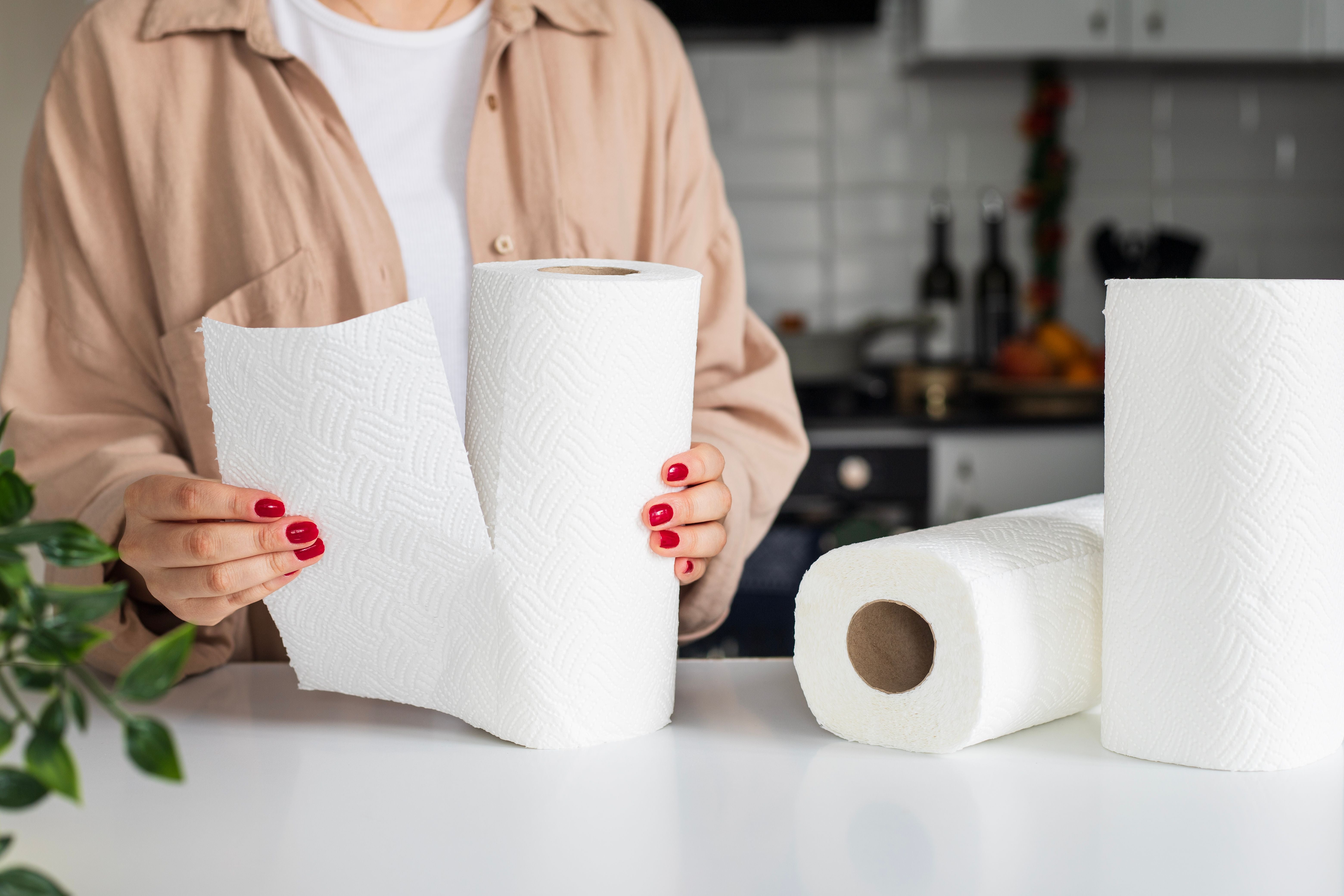Frau reißt Küchenpapier ab, drei Rollen auf weißem Tisch in moderner Küche.