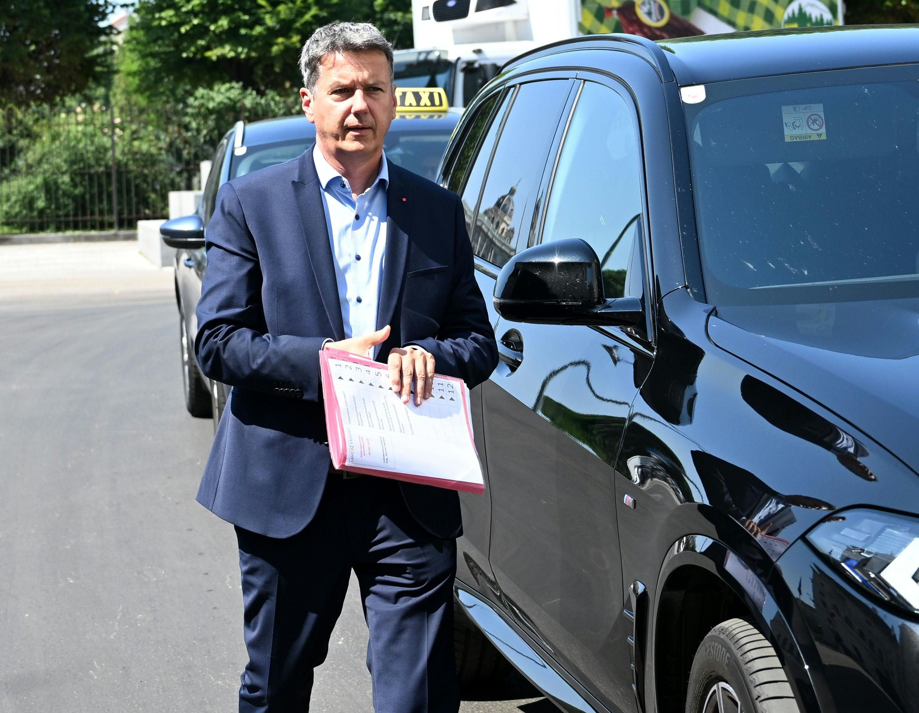 Ein Mann im Anzug hält Unterlagen und steht neben einem schwarzen Auto auf einer Straße.