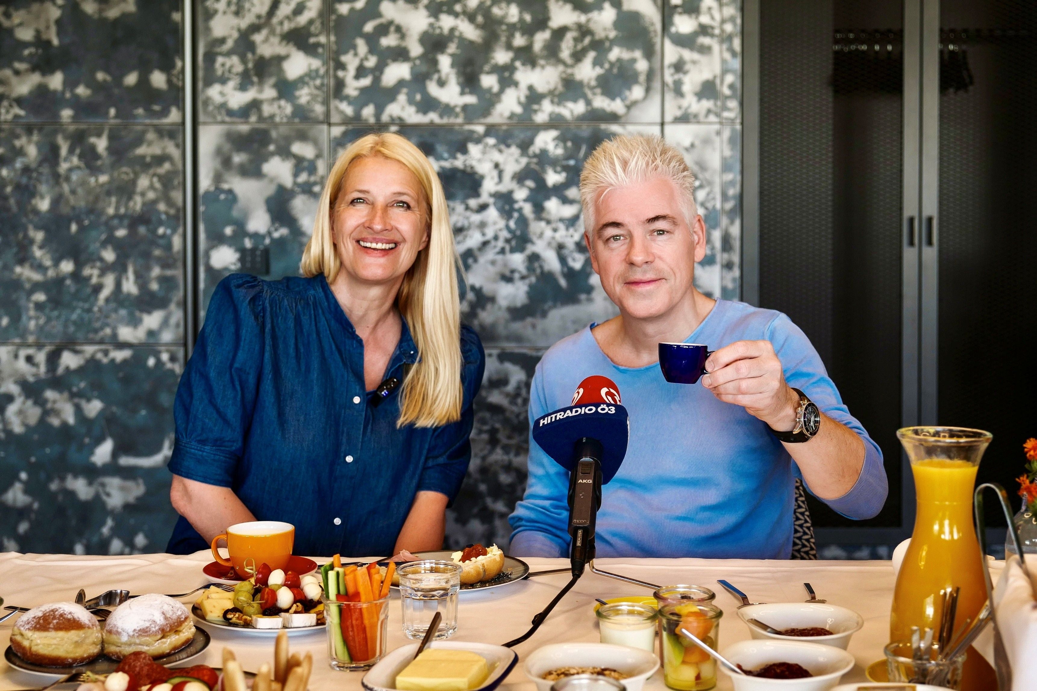 Zwei Menschen sitzen lachend am Frühstückstisch mit Kaffee, Gebäck und Mikrofon.
