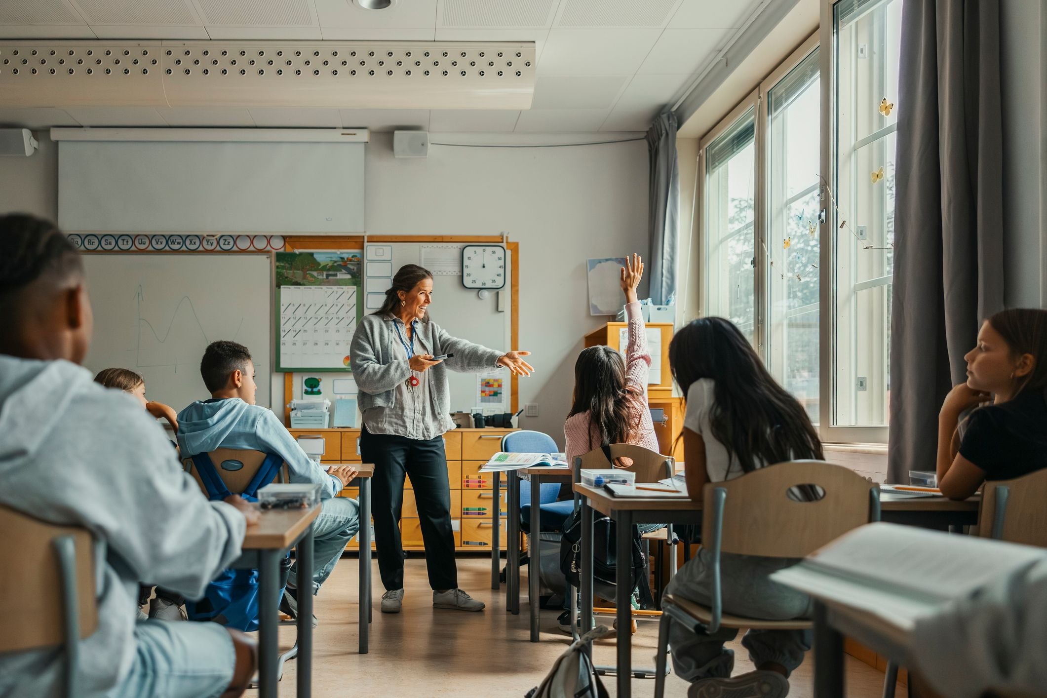 Lehrerin steht vor Schülern im Klassenzimmer, ein Mädchen meldet sich.