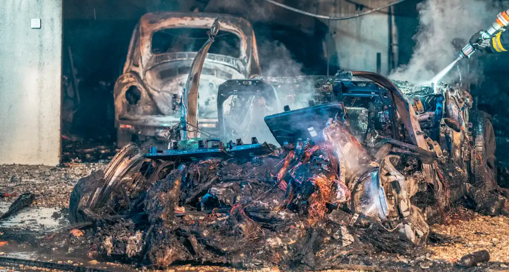 Ausgebrannter E-Jaguar nach Brand, im Hintergrund ein verbranntes Auto, Feuerwehr im Einsatz.