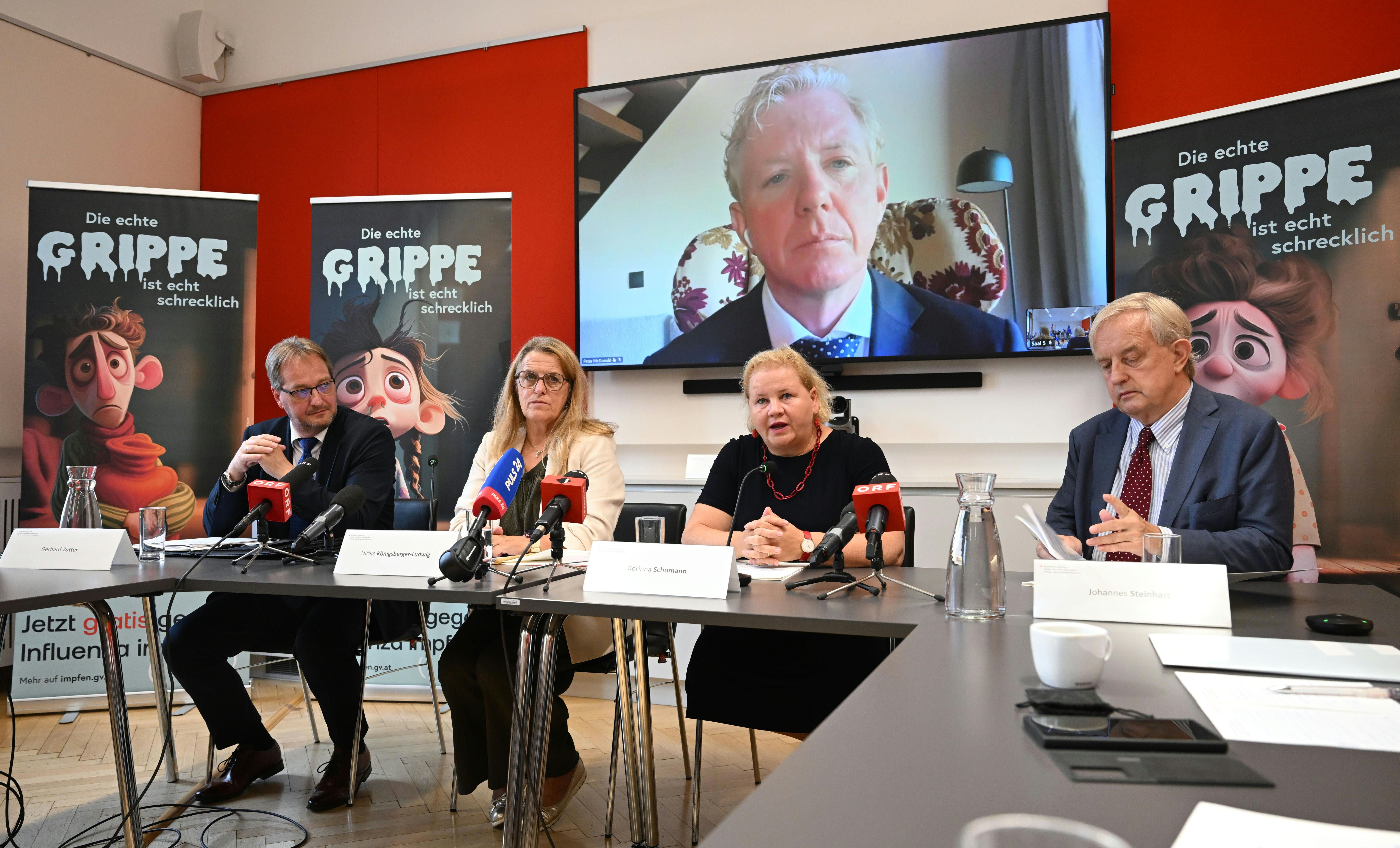 Pressekonferenz mit vier Personen am Tisch, großer Bildschirm und Grippe-Aufklärungsplakate im Hintergrund.
