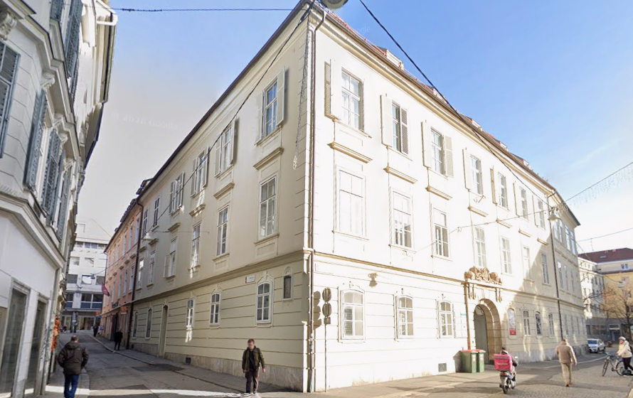 Straßenszene mit historischem Gebäude an einer Ecke und mehreren Fußgängern bei Tageslicht.