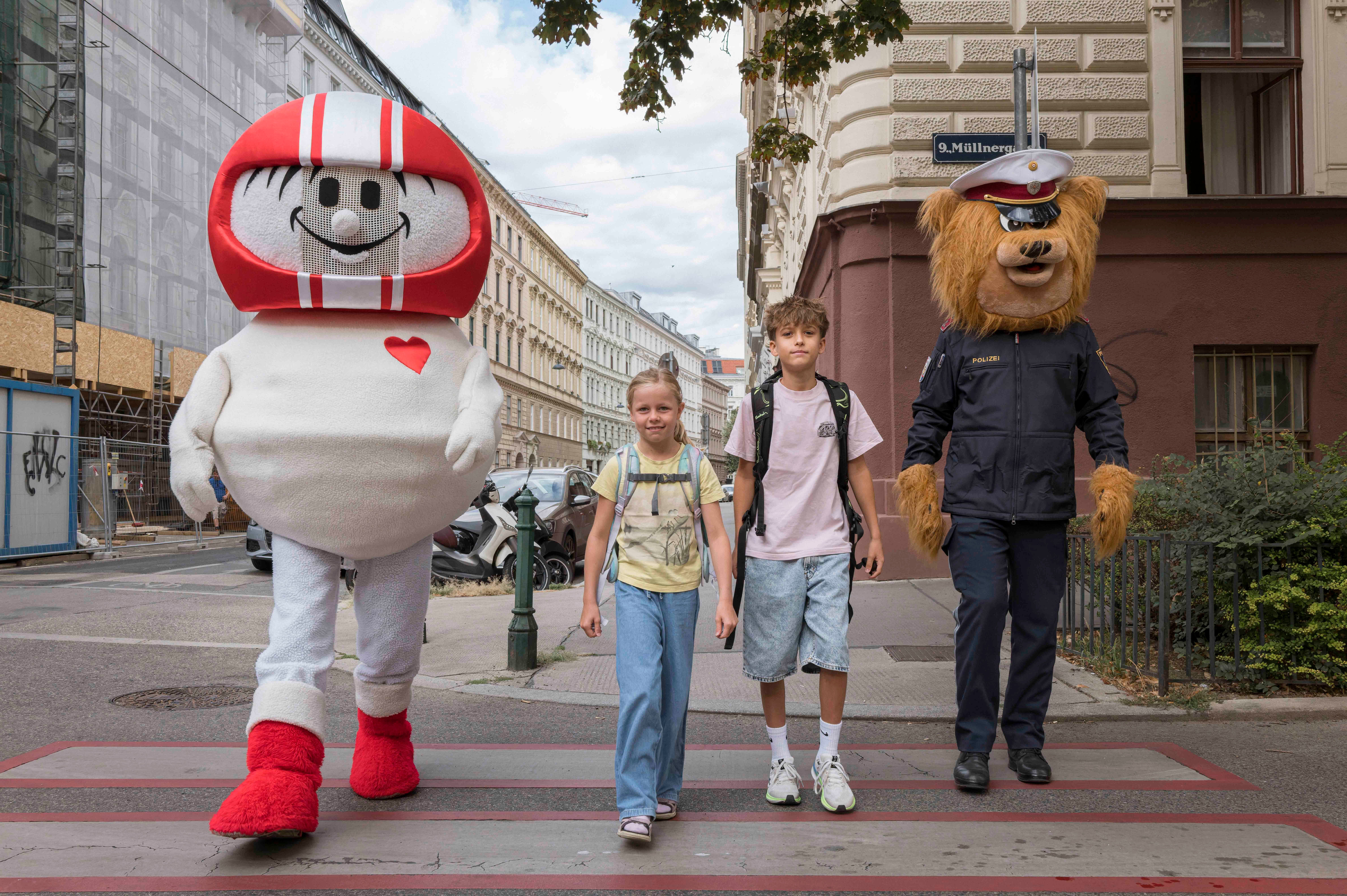 Zwei Kinder überqueren mit Maskottchen einen Zebrastreifen in Wien zum Thema Schulwegsicherheit.