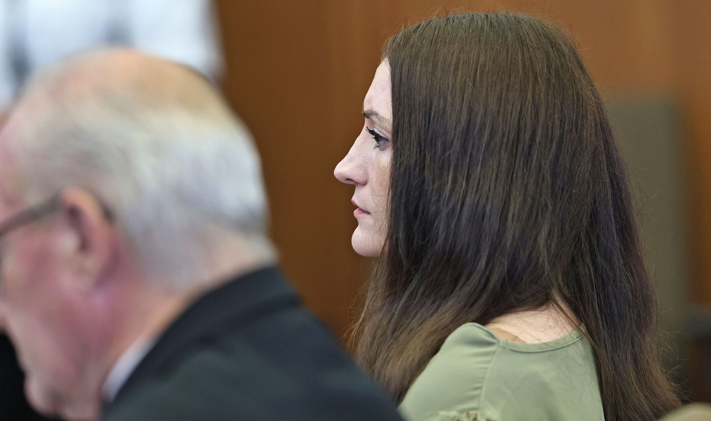 Eine Frau mit langen braunen Haaren sitzt nachdenklich neben einem älteren Mann im Gerichtssaal.