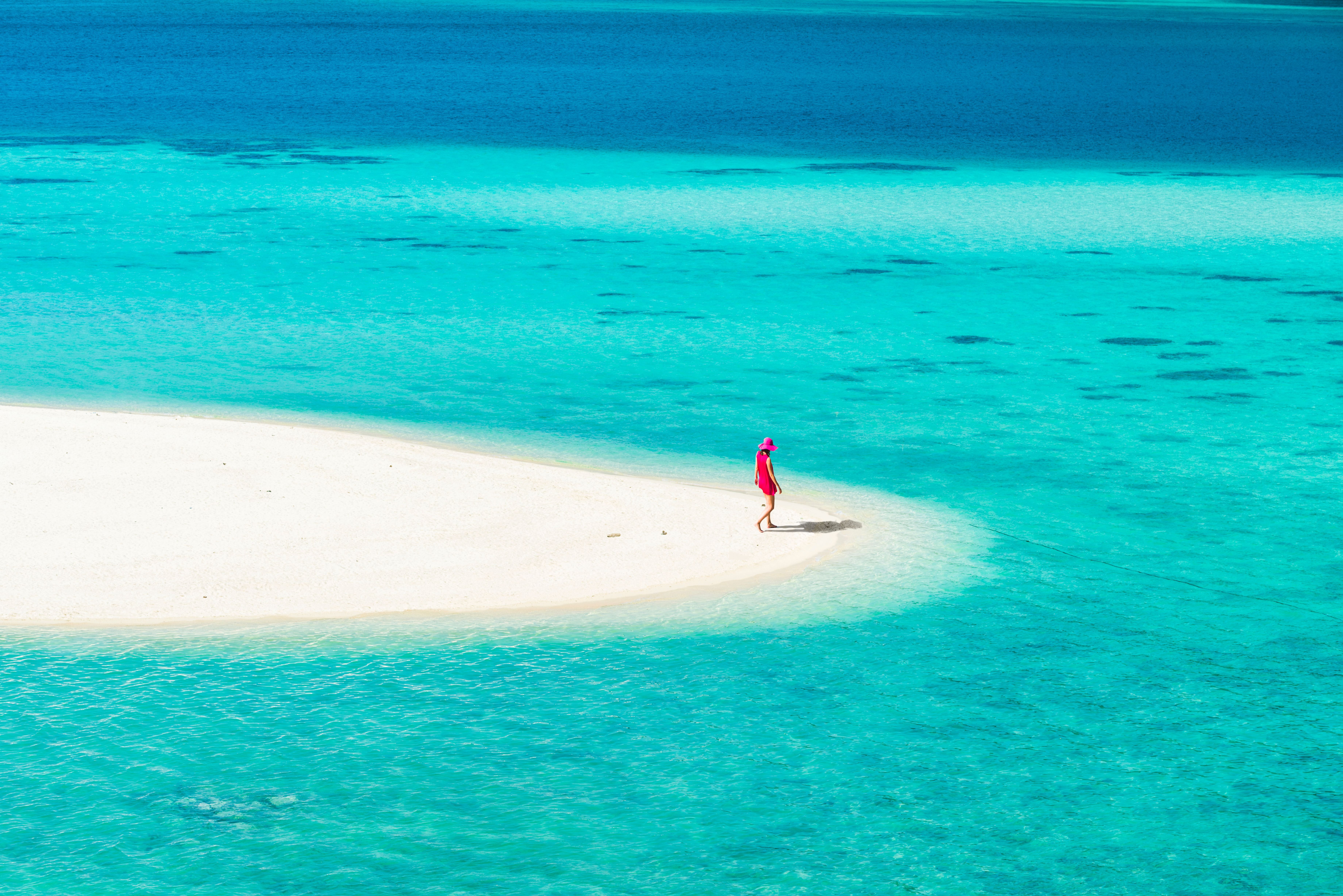 Person mit pinkem Kleid und Hut steht auf einer kleinen Sandinsel im türkisblauen Meer.