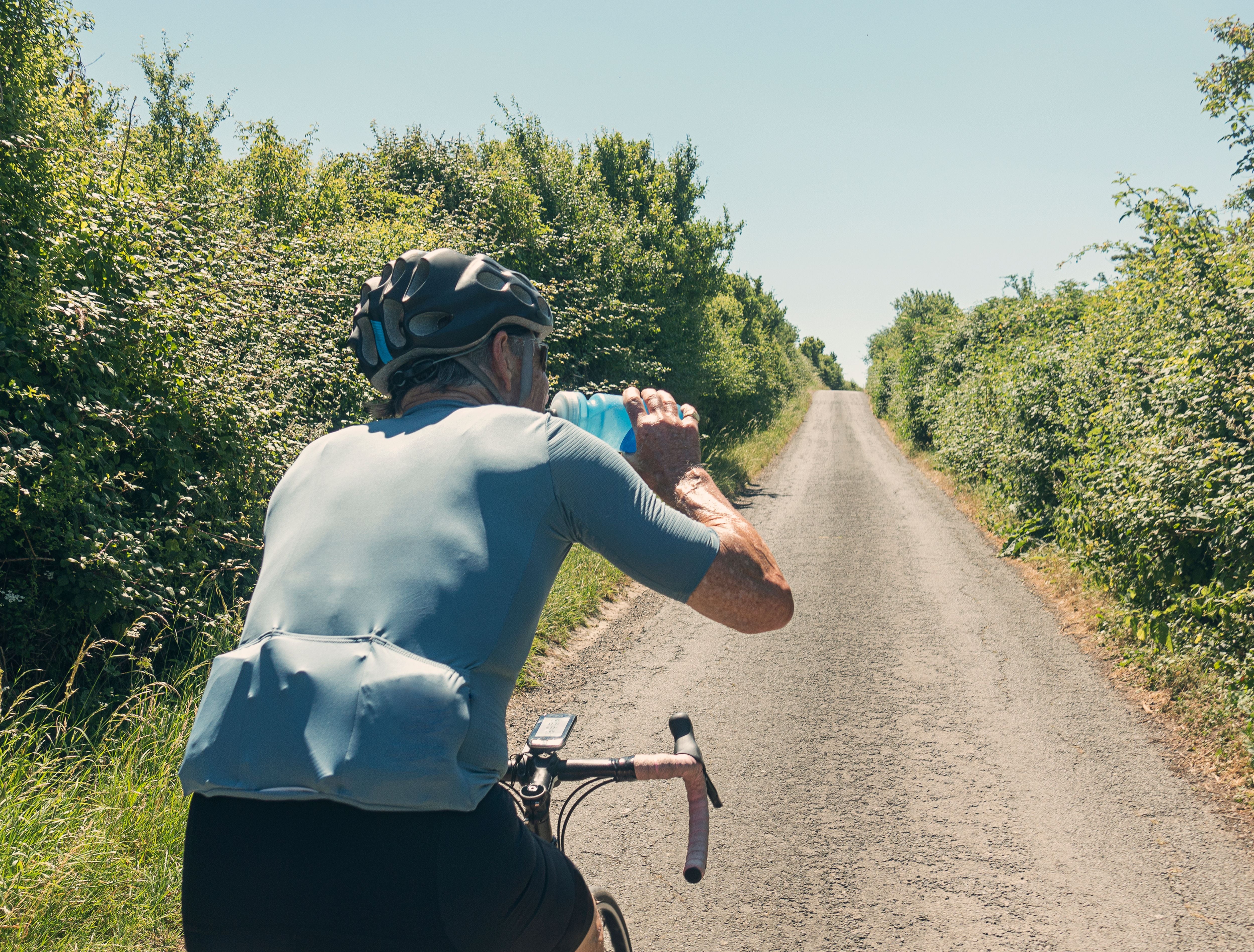 Radfahrer mit Helm trinkt auf einer Landstraße Wasser aus einer Flasche, sonniges Wetter.
