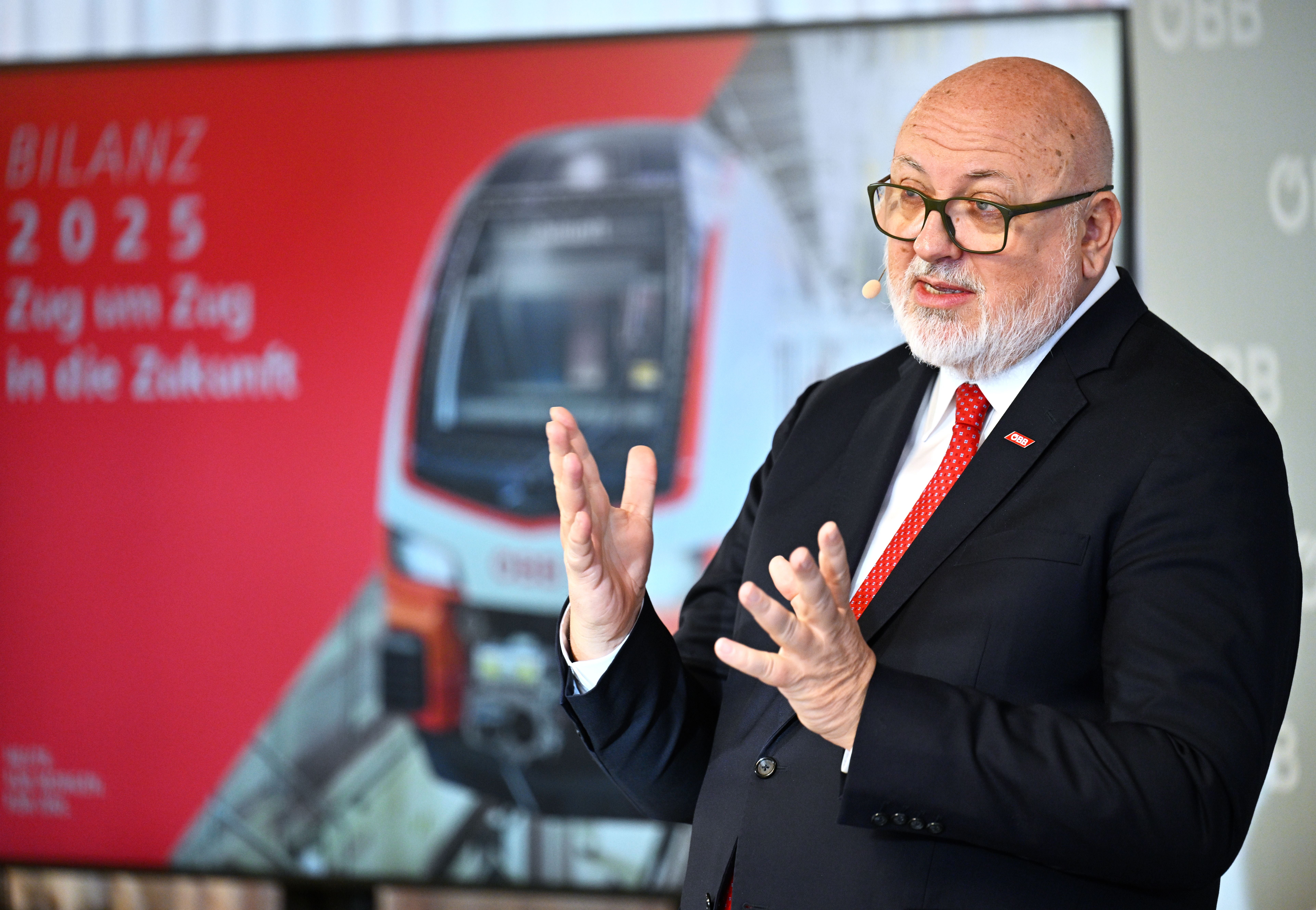 Mann im Anzug spricht bei einer ÖBB-Pressekonferenz, im Hintergrund ein Zug und rotes Plakat.