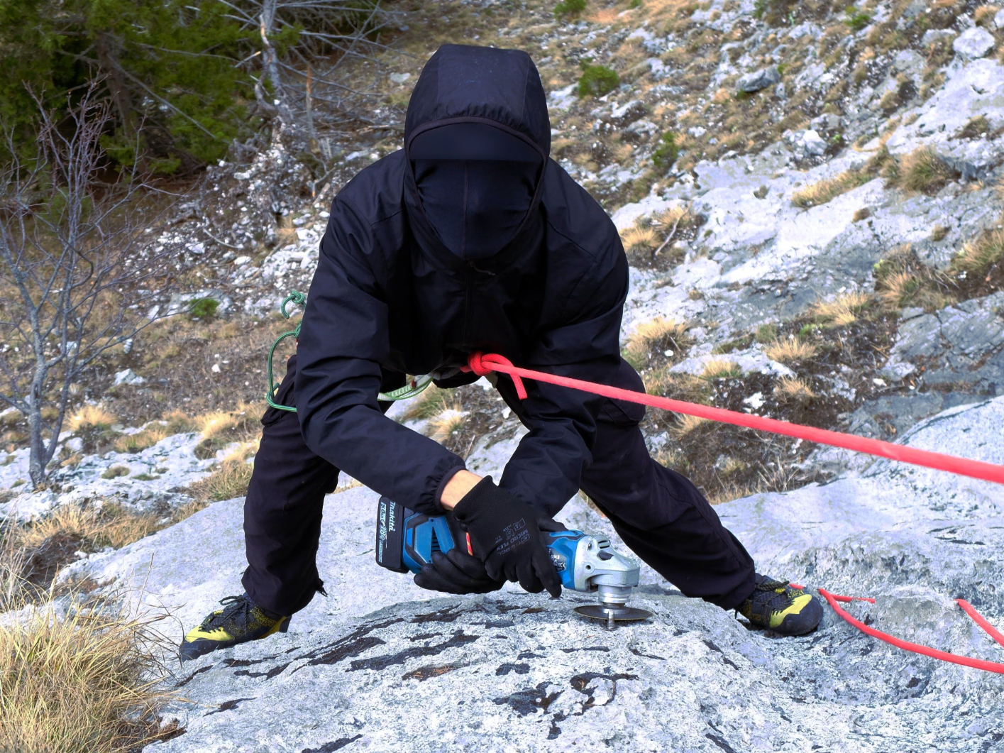 Person in schwarzer Kleidung entfernt mit einer Flex Felshaken im Gebirge, mit Seil gesichert.