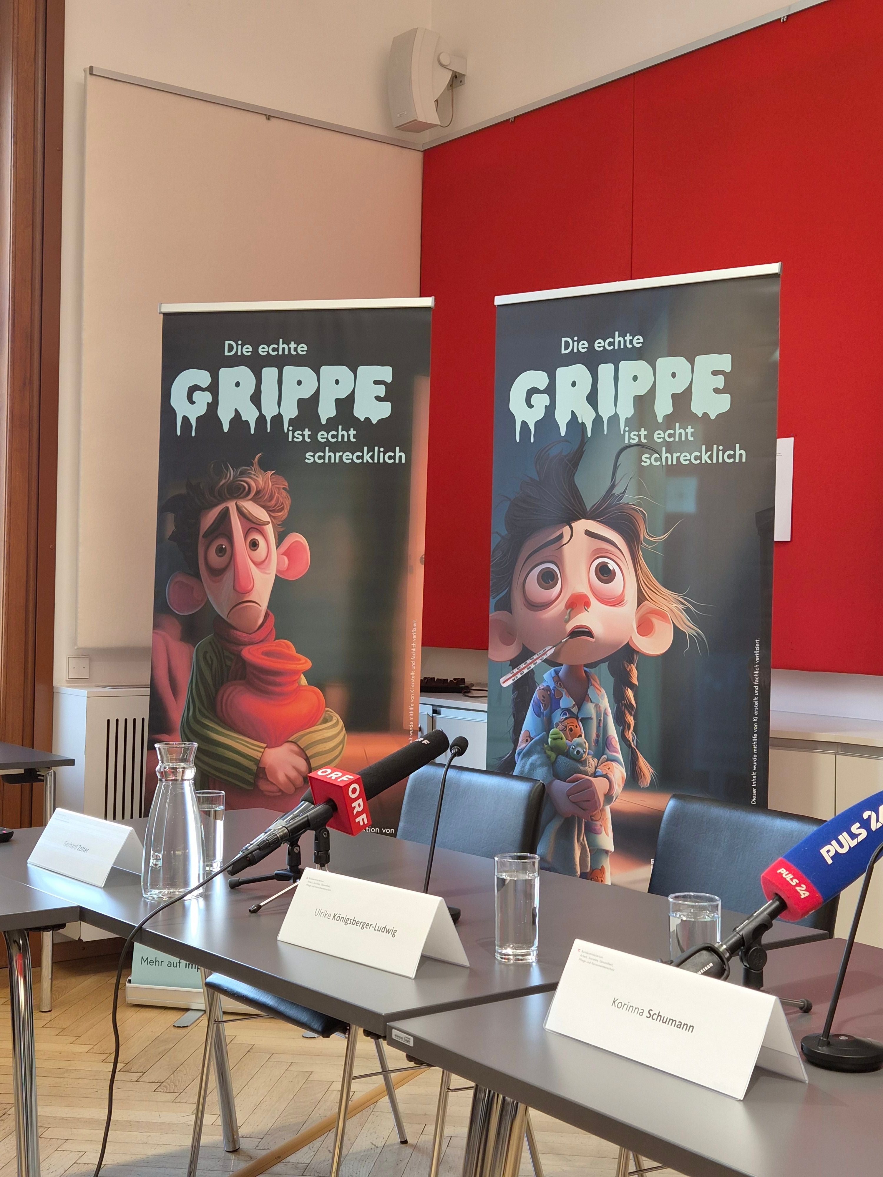 Plakate mit Comicfiguren und dem Text „Die echte Grippe ist echt schrecklich“ bei einer Pressekonferenz.