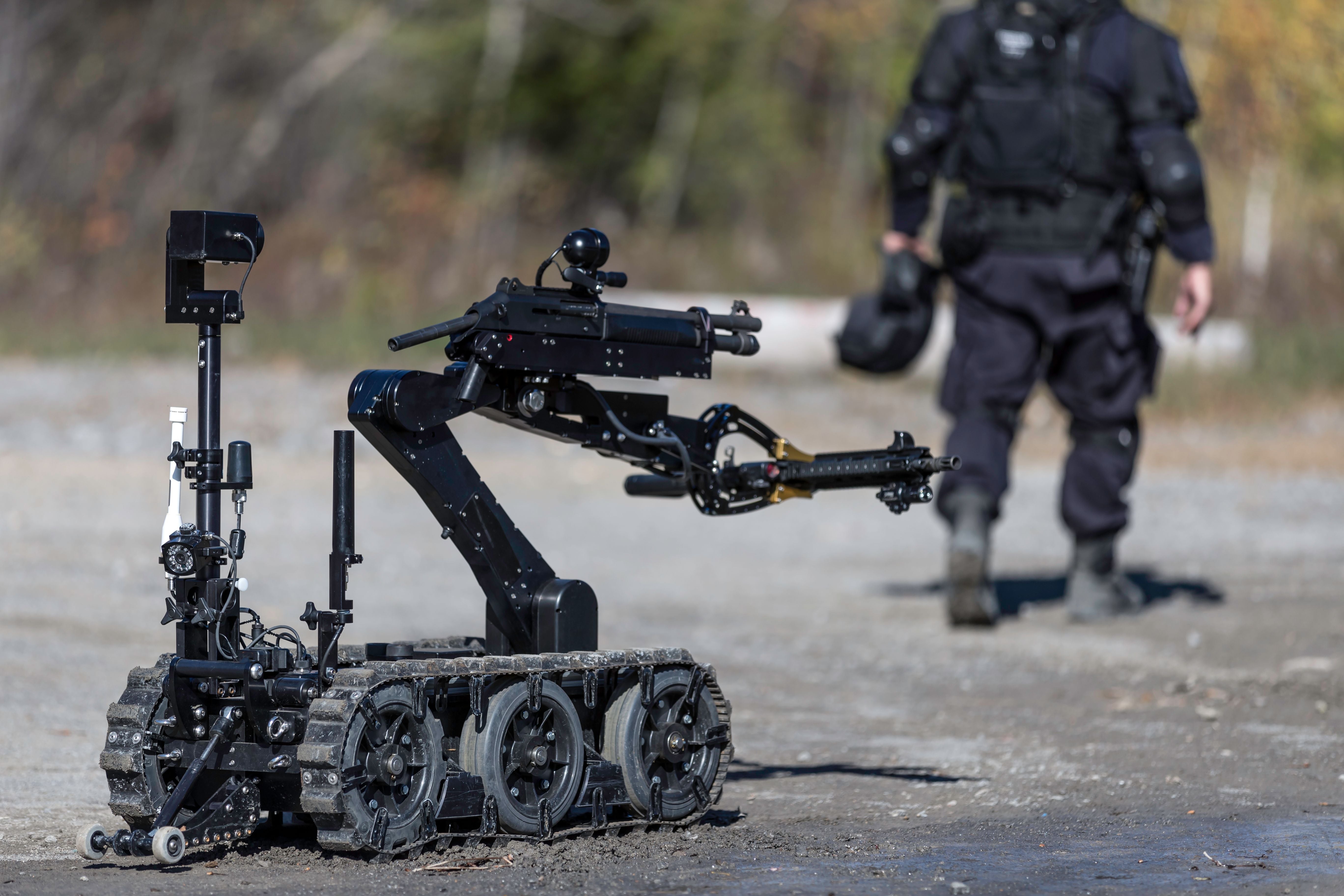Ein Bombenentschärfungsroboter mit Greifarm steht vor einem Polizisten in Schutzausrüstung.