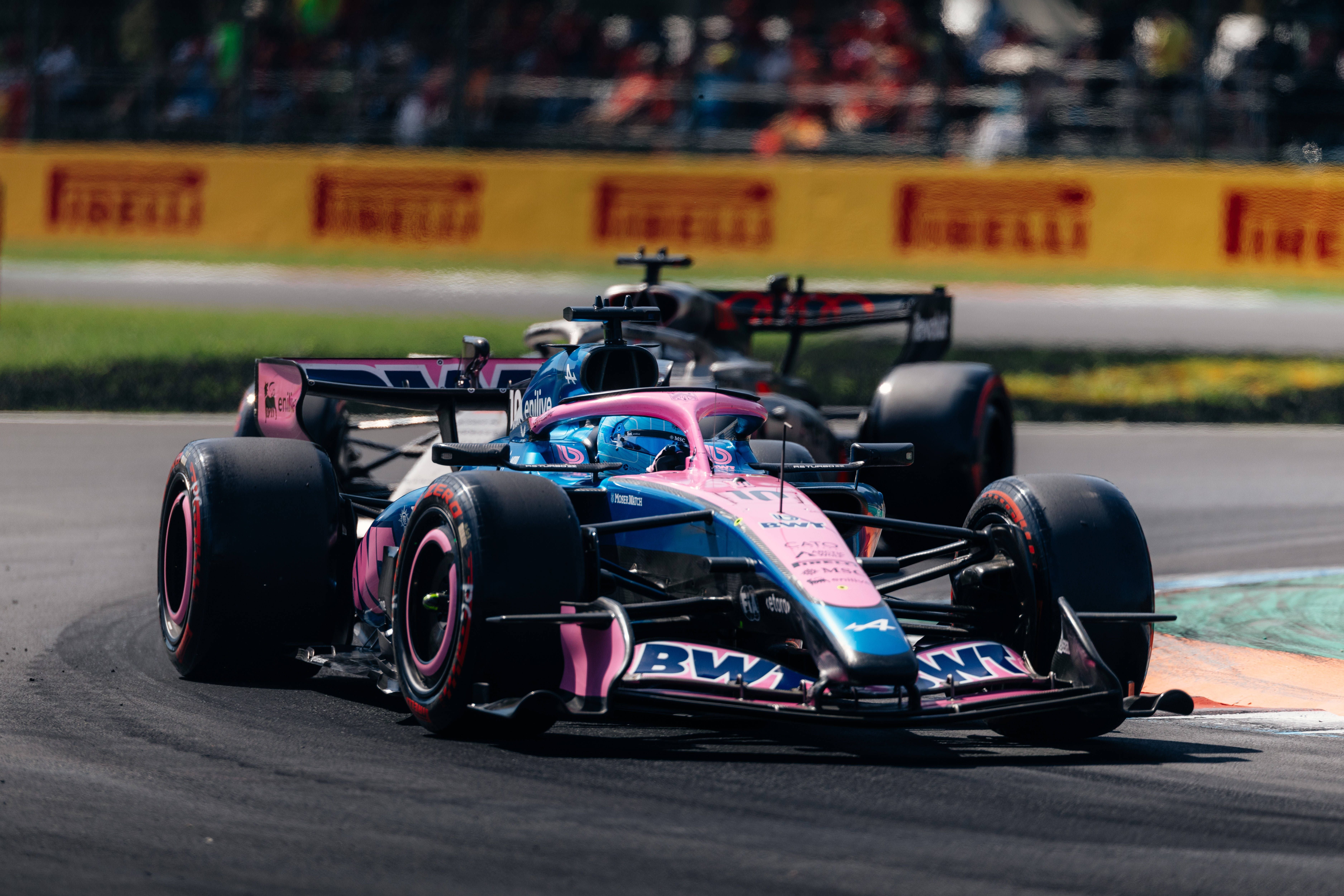 Ein pink-blauer Formel-1-Wagen fährt vor einem anderen Auto durch eine Kurve auf einer Rennstrecke.