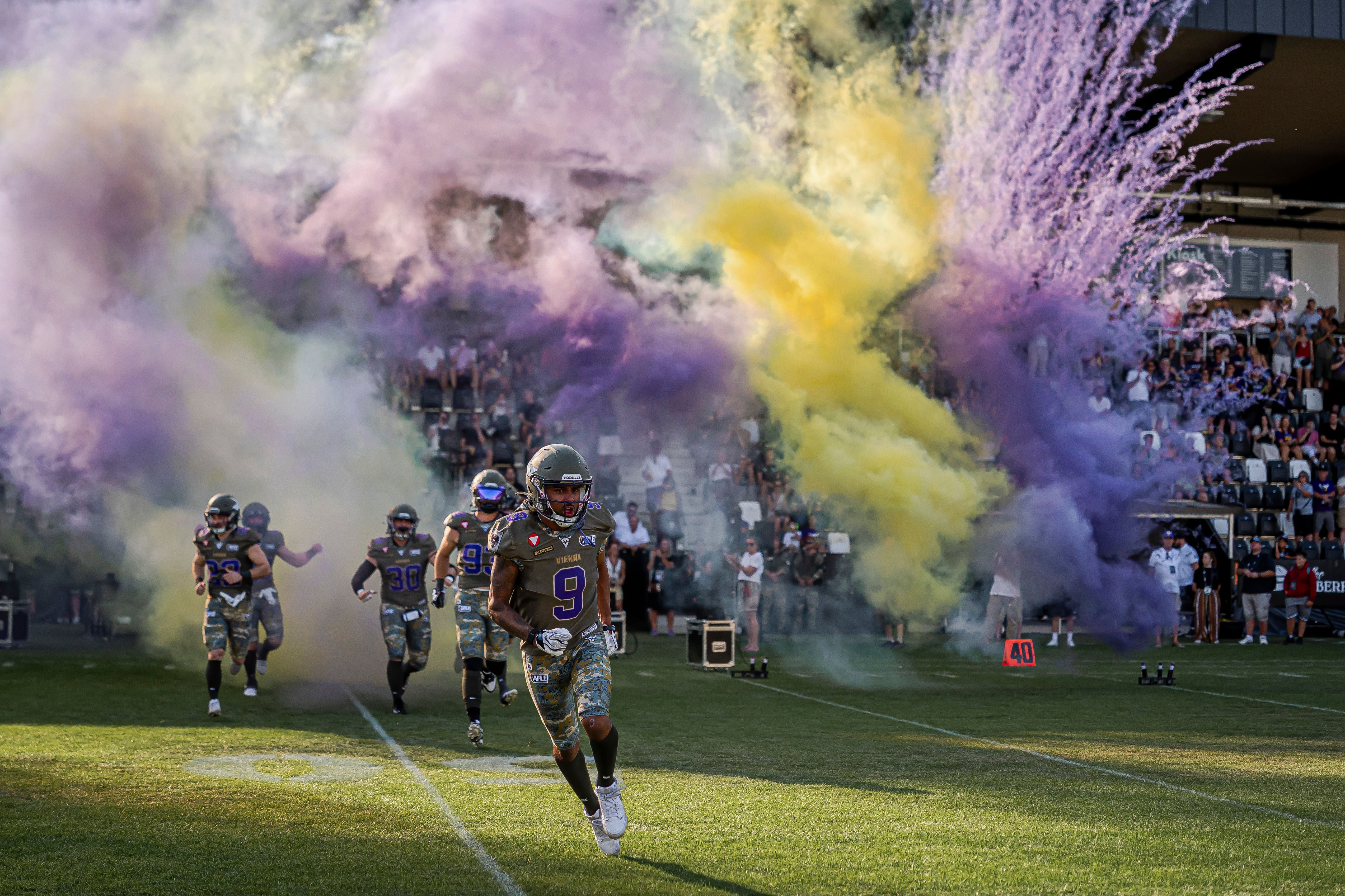 Footballspieler der Vikings laufen durch lila und gelben Rauch aufs Spielfeld.