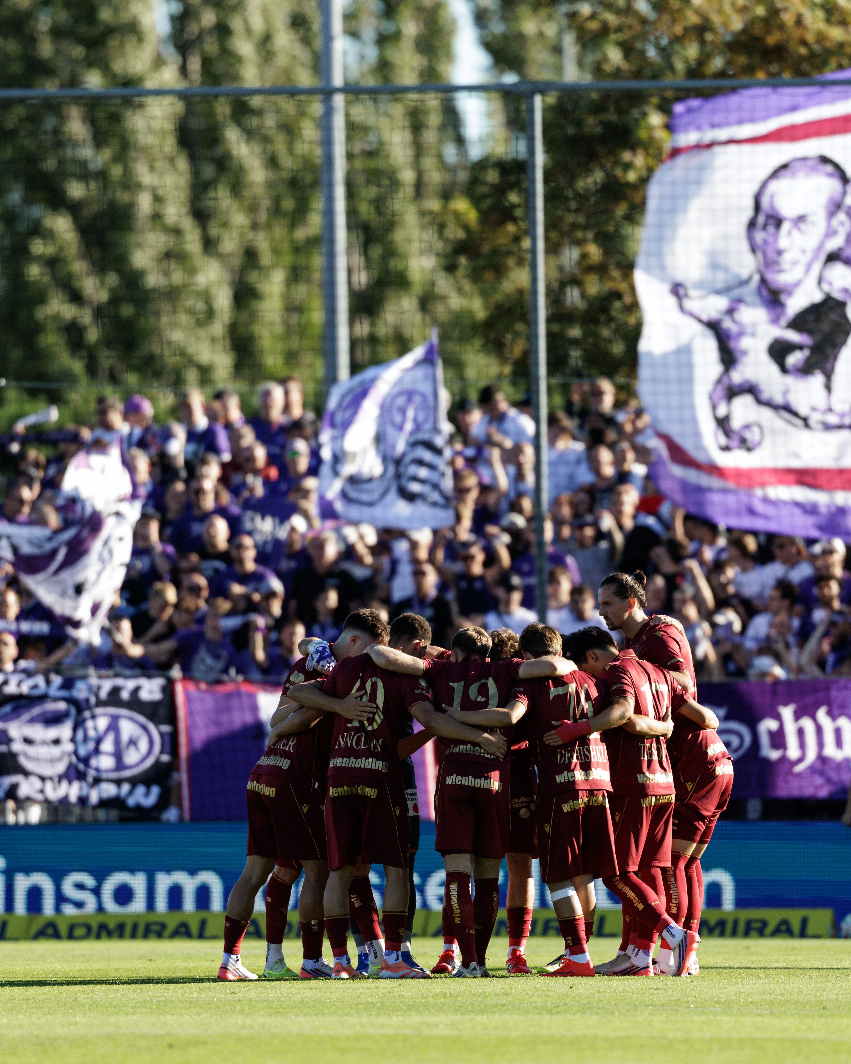 Eine Fußballmannschaft in roten Trikots steht im Kreis vor jubelnden Fans mit violetten Bannern.
