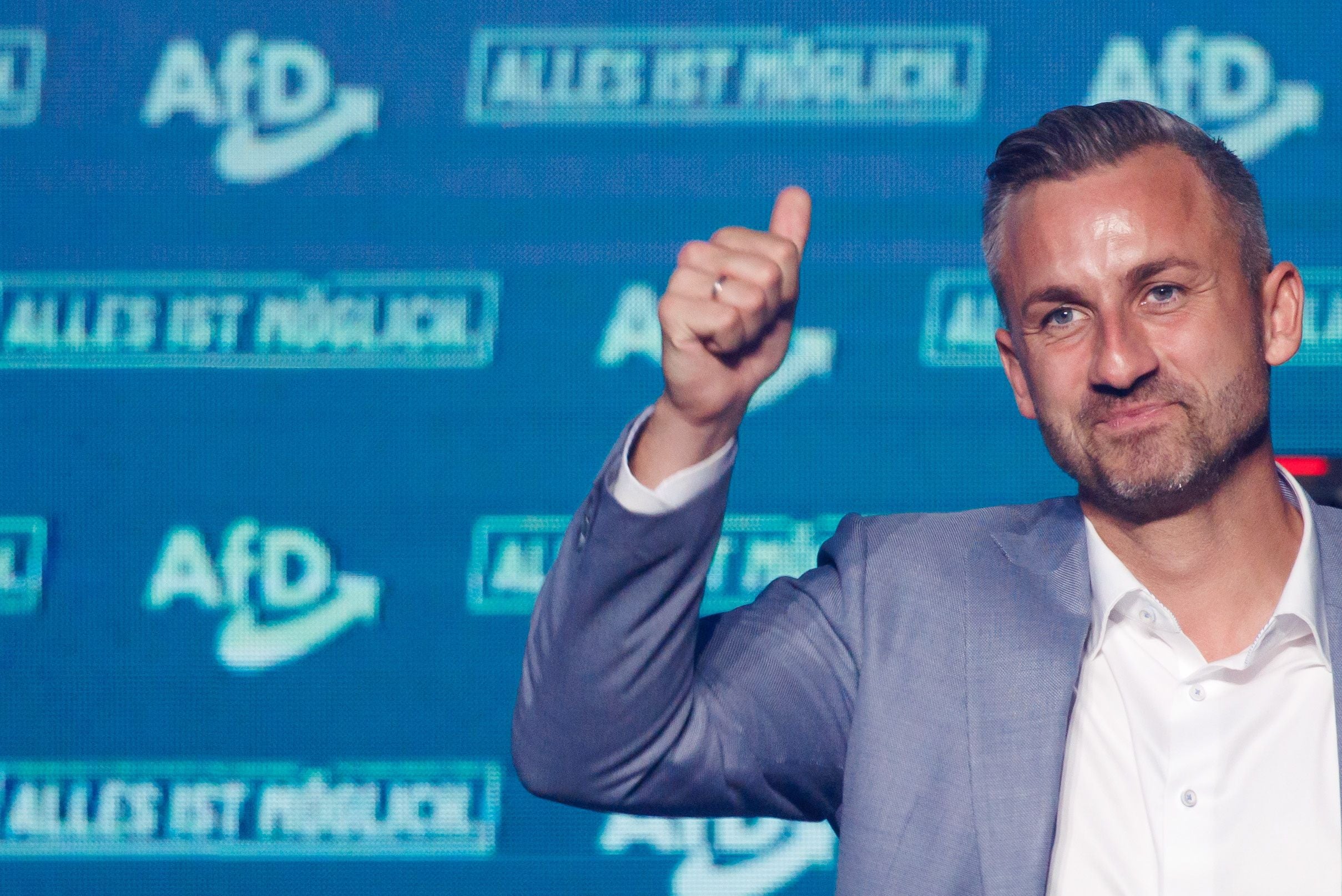 Ein Mann im Anzug zeigt Daumen hoch vor einer AfD-Wand bei einer Wahlkampfveranstaltung.