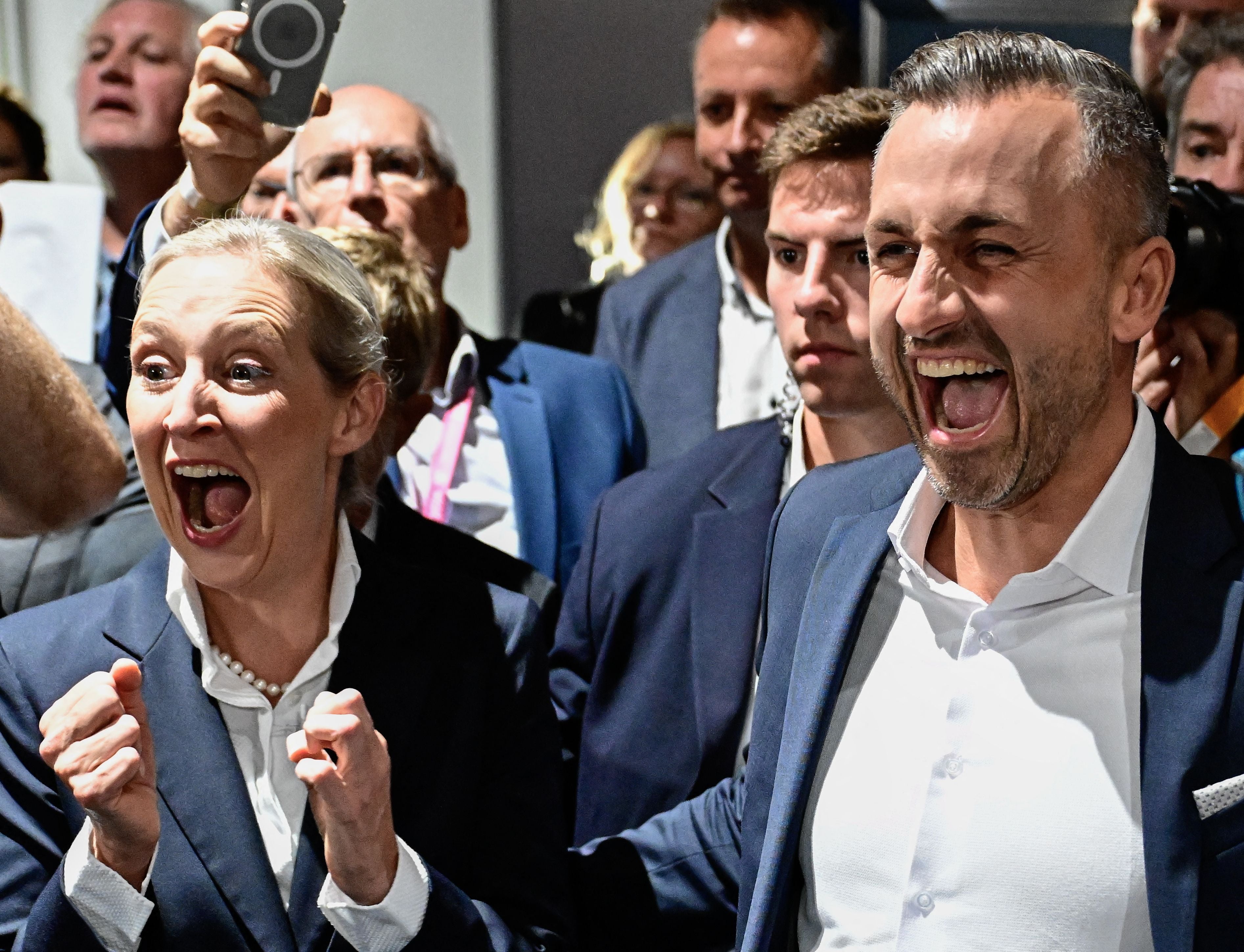 Zwei Menschen im Anzug jubeln lautstark in einer Menschenmenge bei einer Wahlveranstaltung.