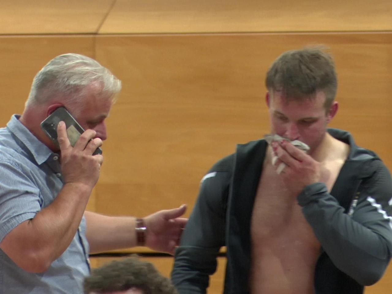 Der verletzte Judoka Timo Cavelius presst ein Tuch vor seinen blutenden Mund, während ein Mann im hemdblauen Oberteil telefonierend neben ihm steht.