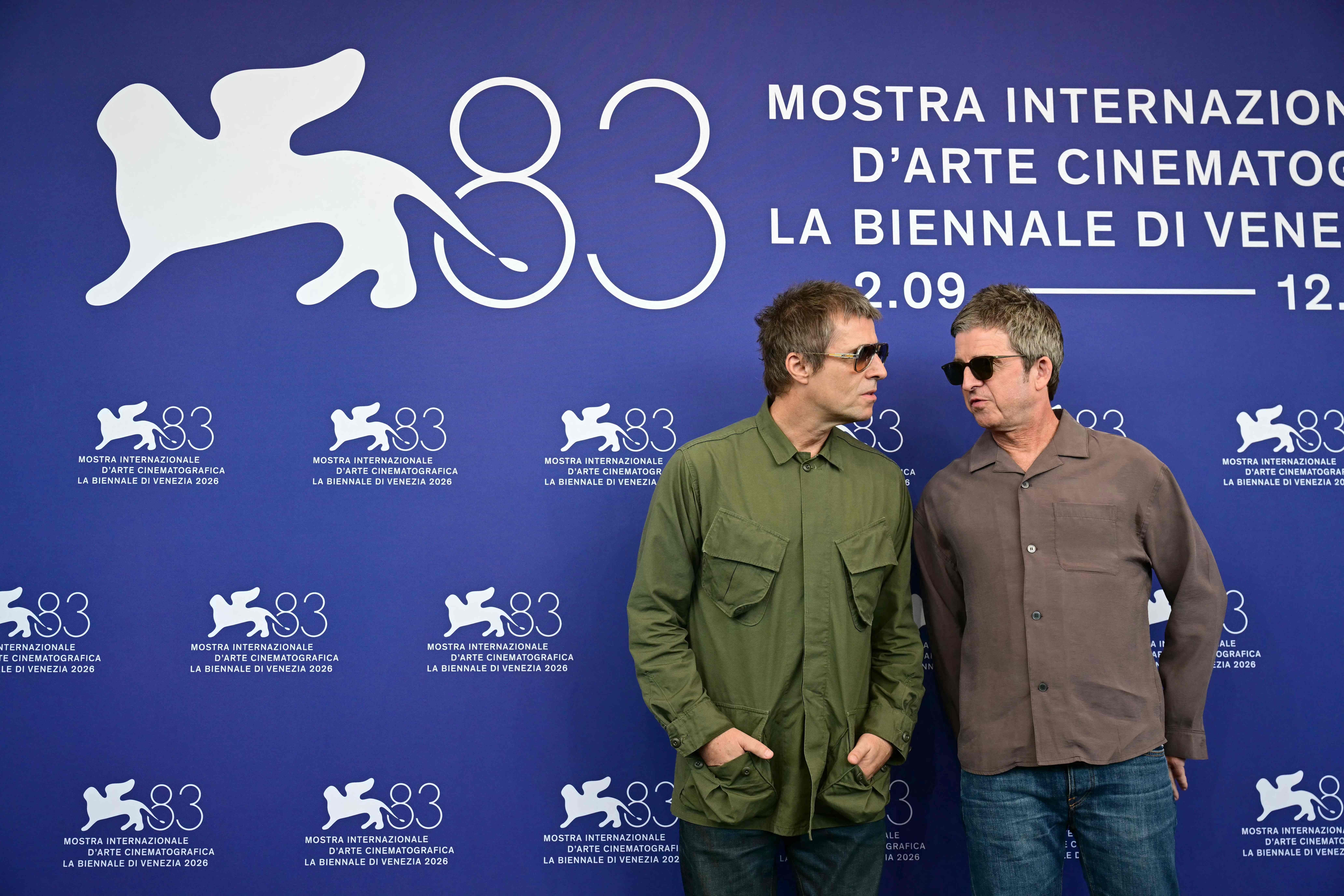 Zwei Männer mit Sonnenbrillen stehen vor einer blauen Wand der 83. Filmfestspiele von Venedig.