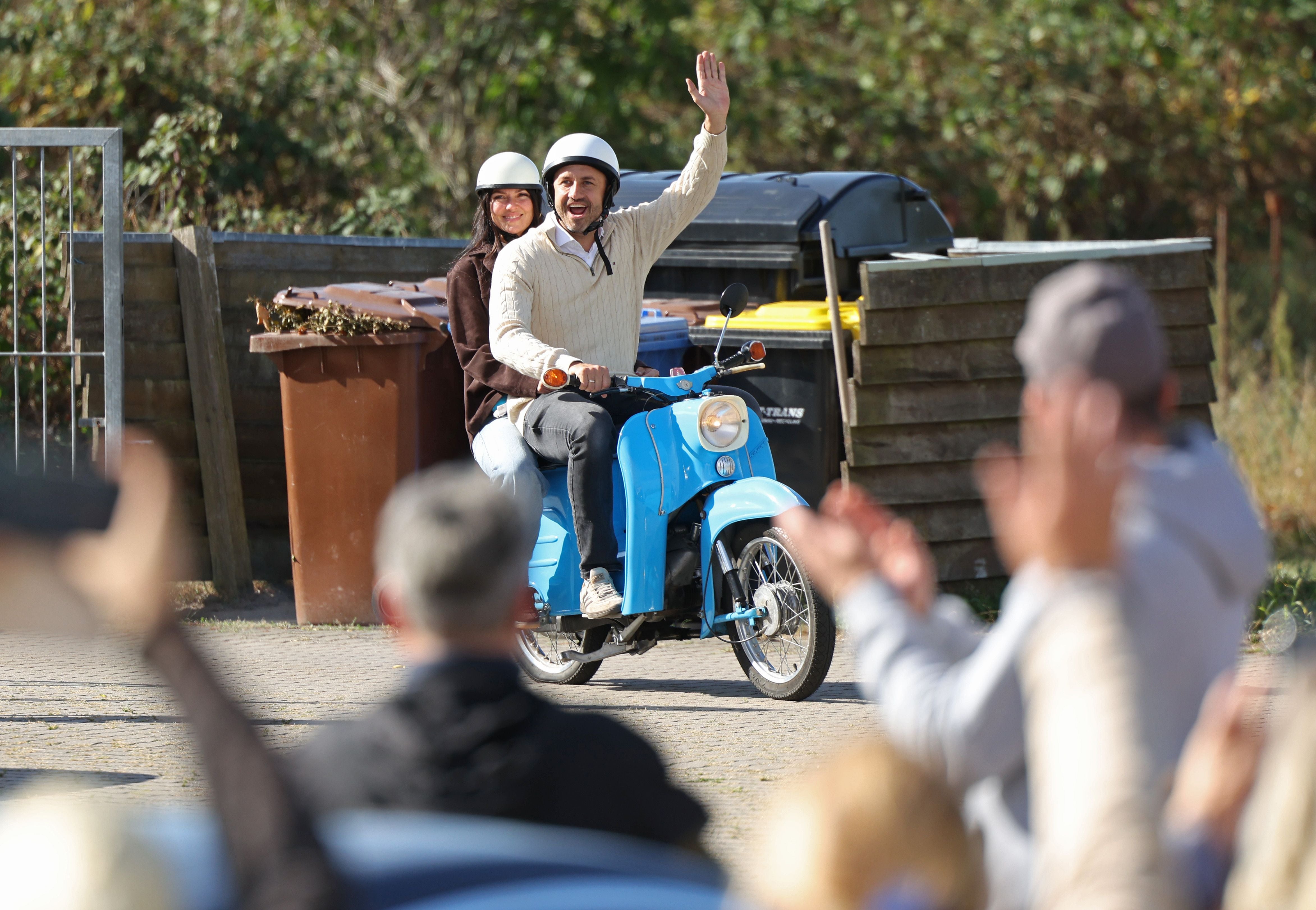 Ein Mann und eine Frau fahren auf einem blauen Moped, der Mann winkt einer applaudierenden Menge.