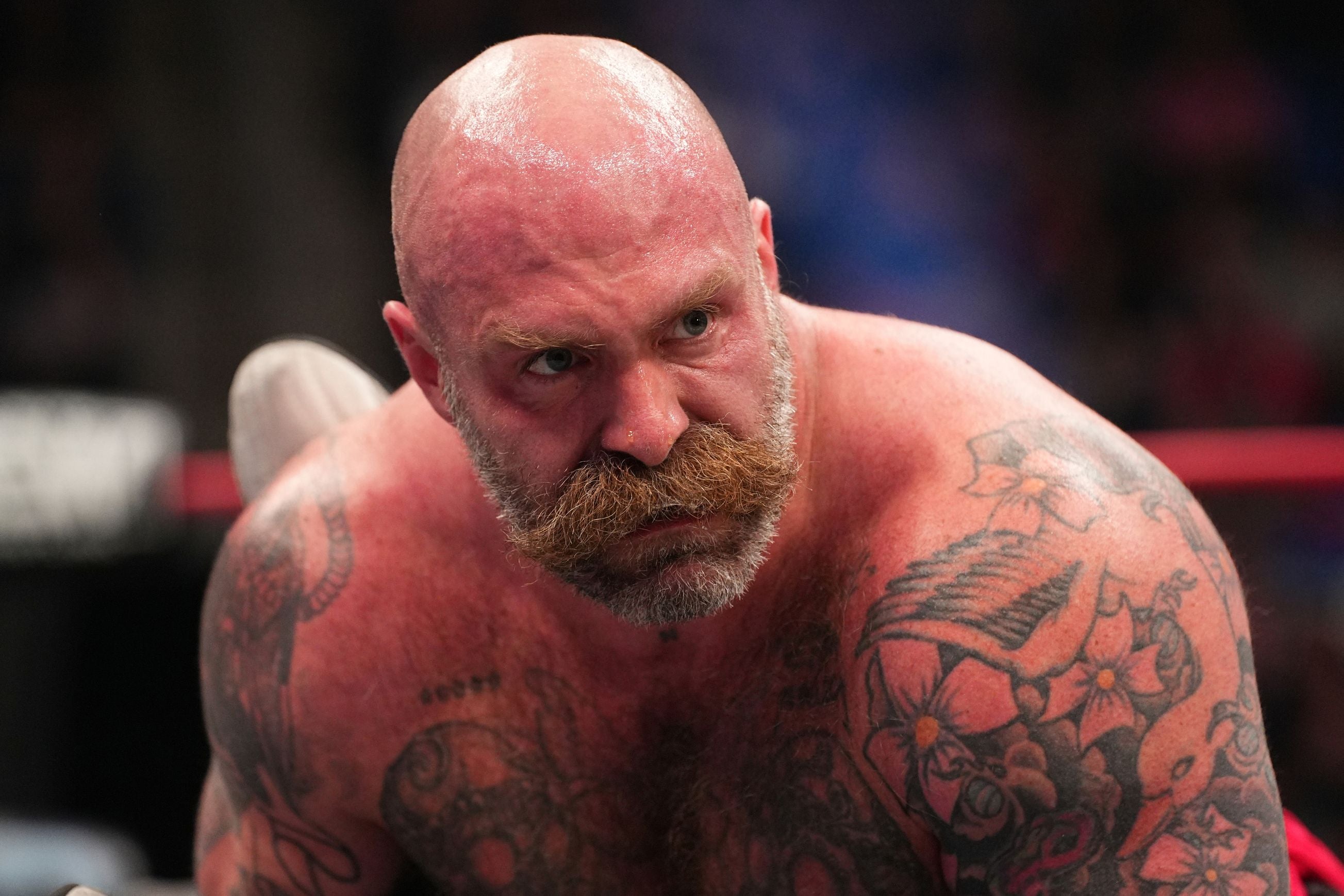 Ein glatzköpfiger, muskulöser Mann mit Bart und Tattoos schaut entschlossen in die Kamera.